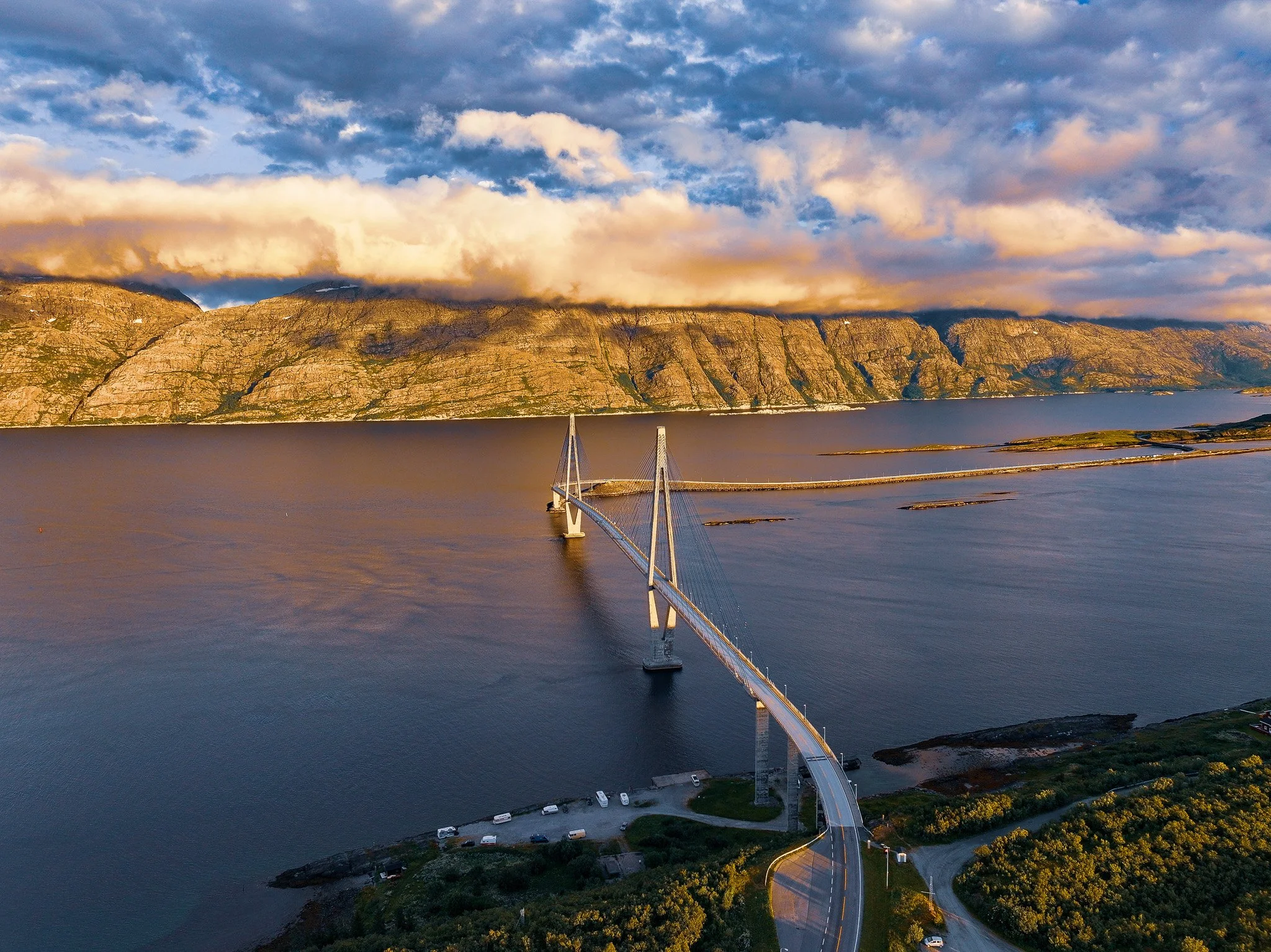 Ein Brückenbauwerk über eine große Wasserfläche, vor einer Berglandschaft, bei Sonnenuntergang mit bewölktem Himmel.