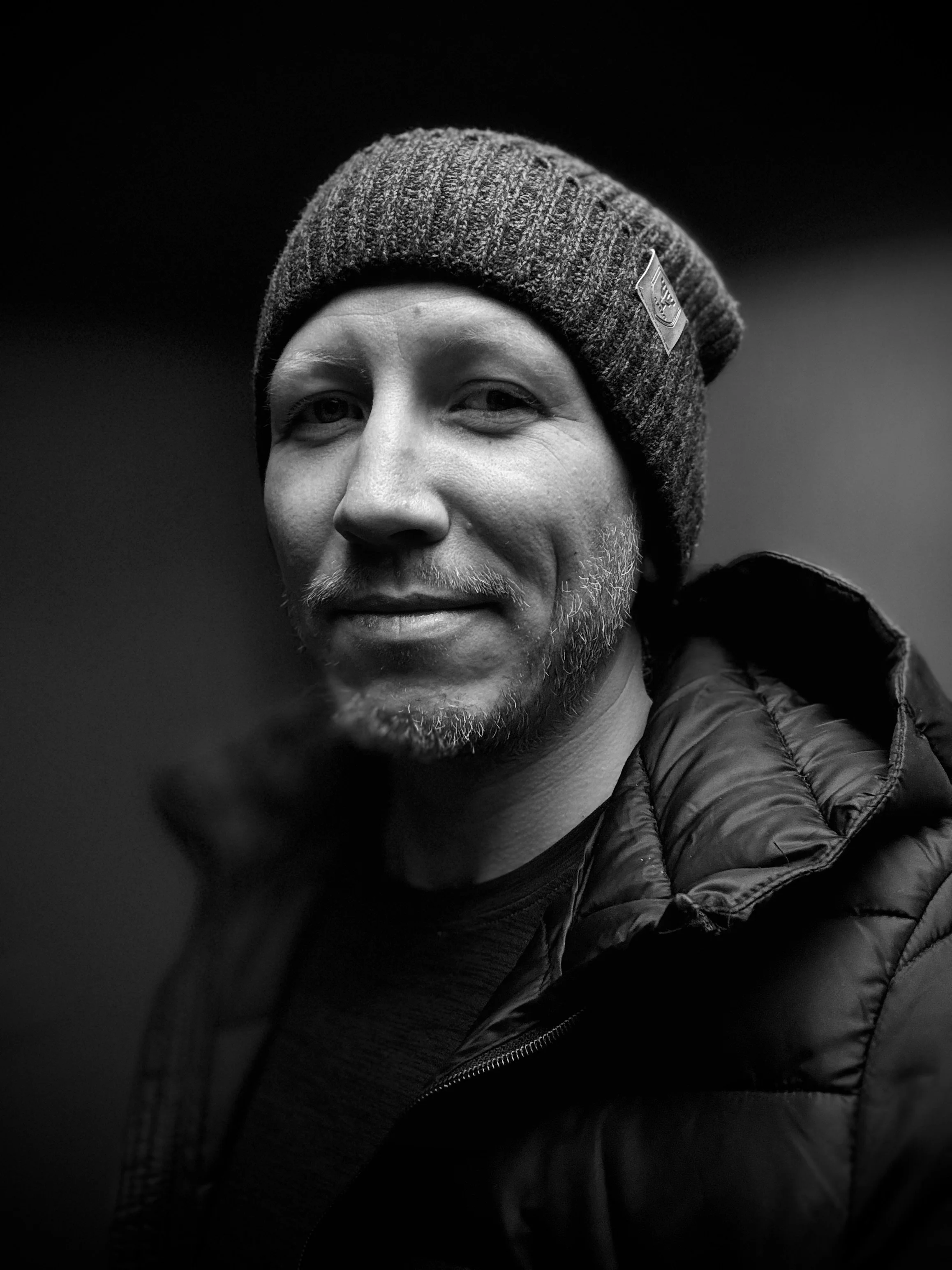 Schwarz-weiße Nahaufnahme eines Mannes mit Mütze und Jacke, lächelnd.