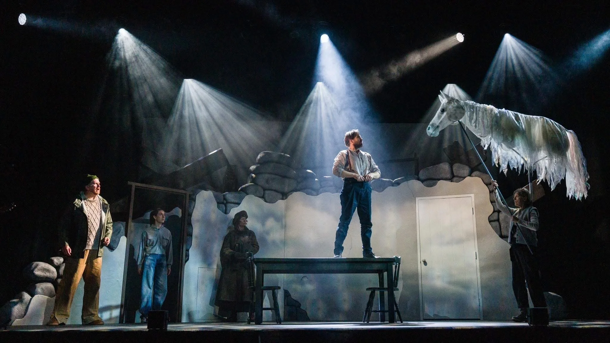 Stage scene with five actors; one standing on a desk, another holding a white flag with a horse head, and three others observing; set resembles a rocky outdoor environment with stage lighting.