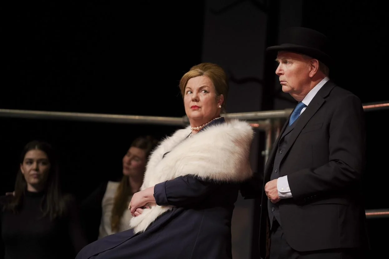 Actors performing in a stage play, with a woman in a dark dress and a white fur stole, and a man in a dark suit and hat, both looking serious.