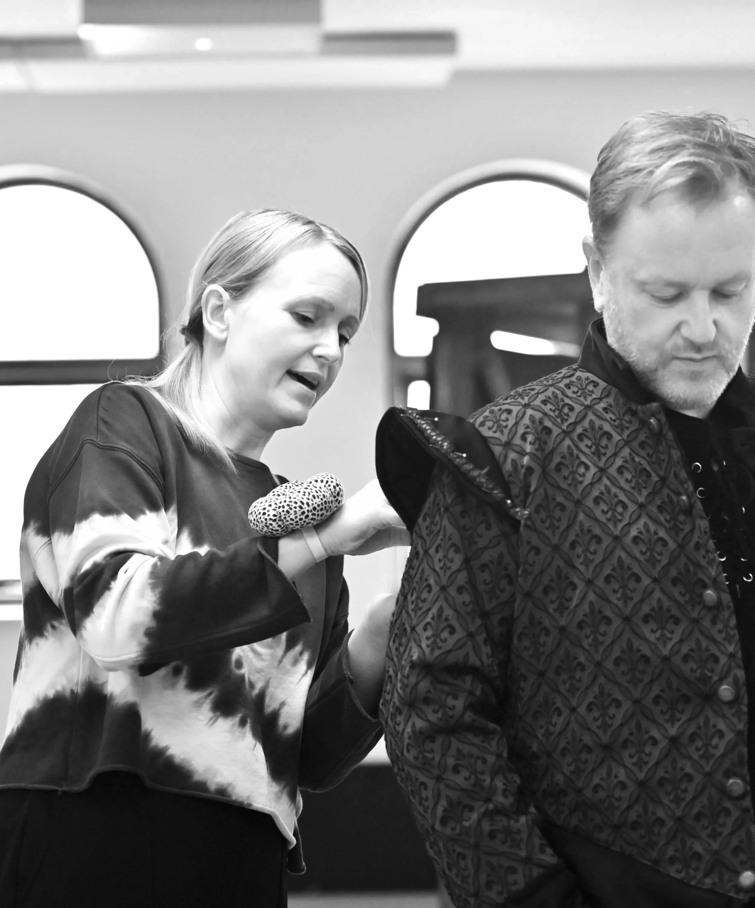 A woman is helping a man with his backpack in a room with arched windows.