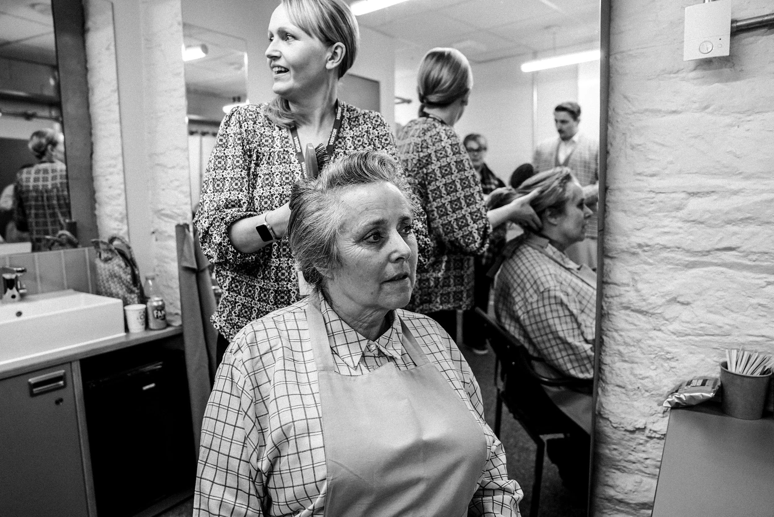 Women getting haircuts in a salon, with a woman seated having her hair styled while another woman stands behind her, and more women getting hair treatments in the background.