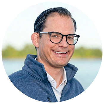 A smiling man with dark glasses and dark hair, wearing a blue jacket outdoors.
