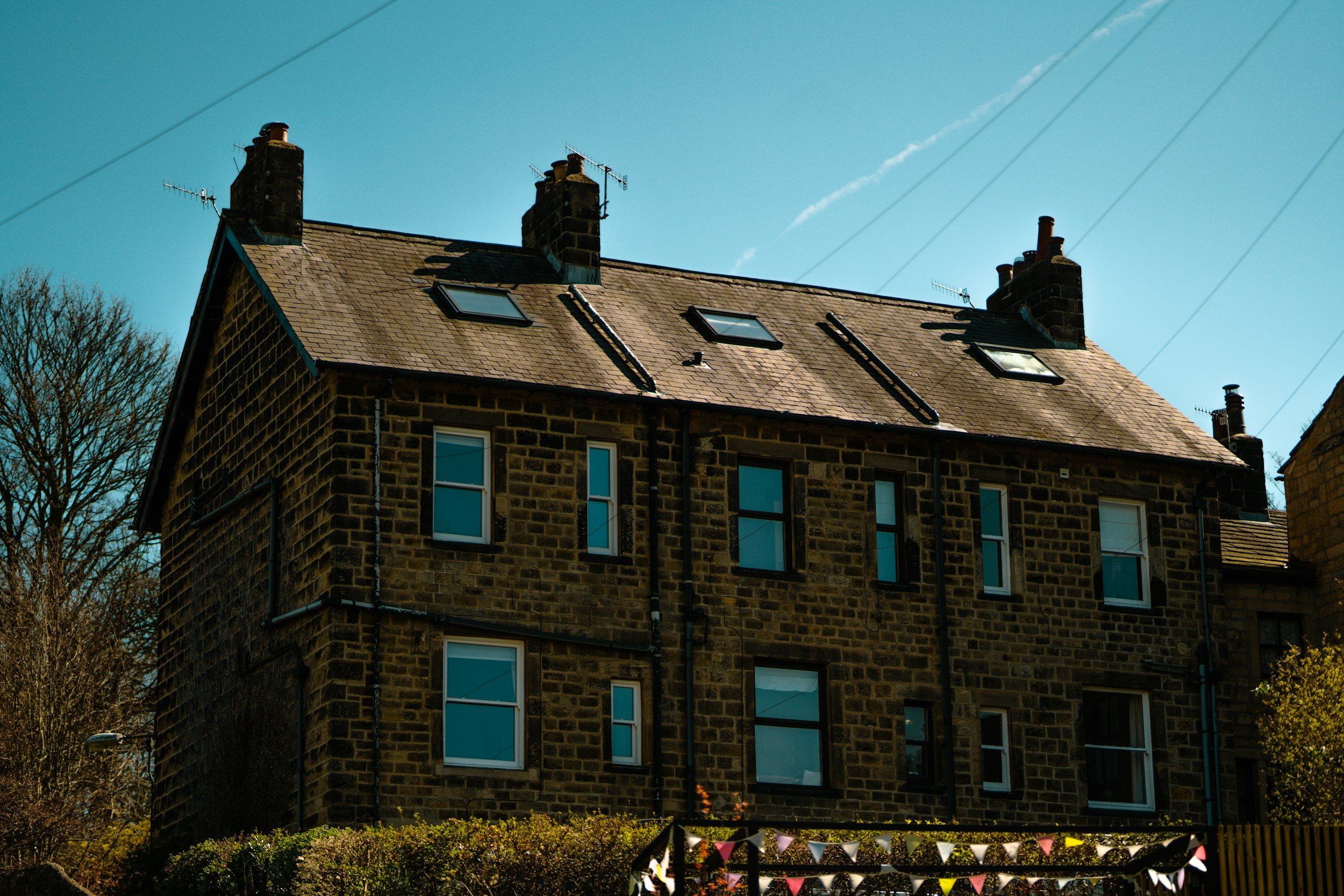 A multi-story brick building with a slate roof, several chimney stacks, and multiple skylights. There are windows on each floor and trees in the background, with power lines overhead.
