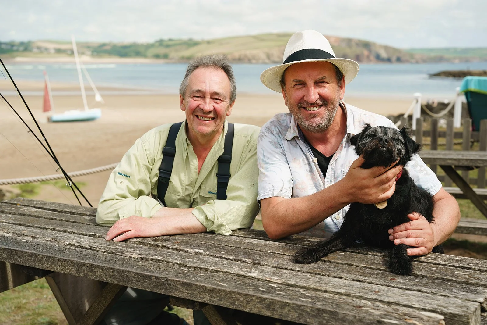 Two smiling men, one with a white hat, sitting at a wooden picnic table by the beach, with a man holding a black dog, and sailboats on the sand.