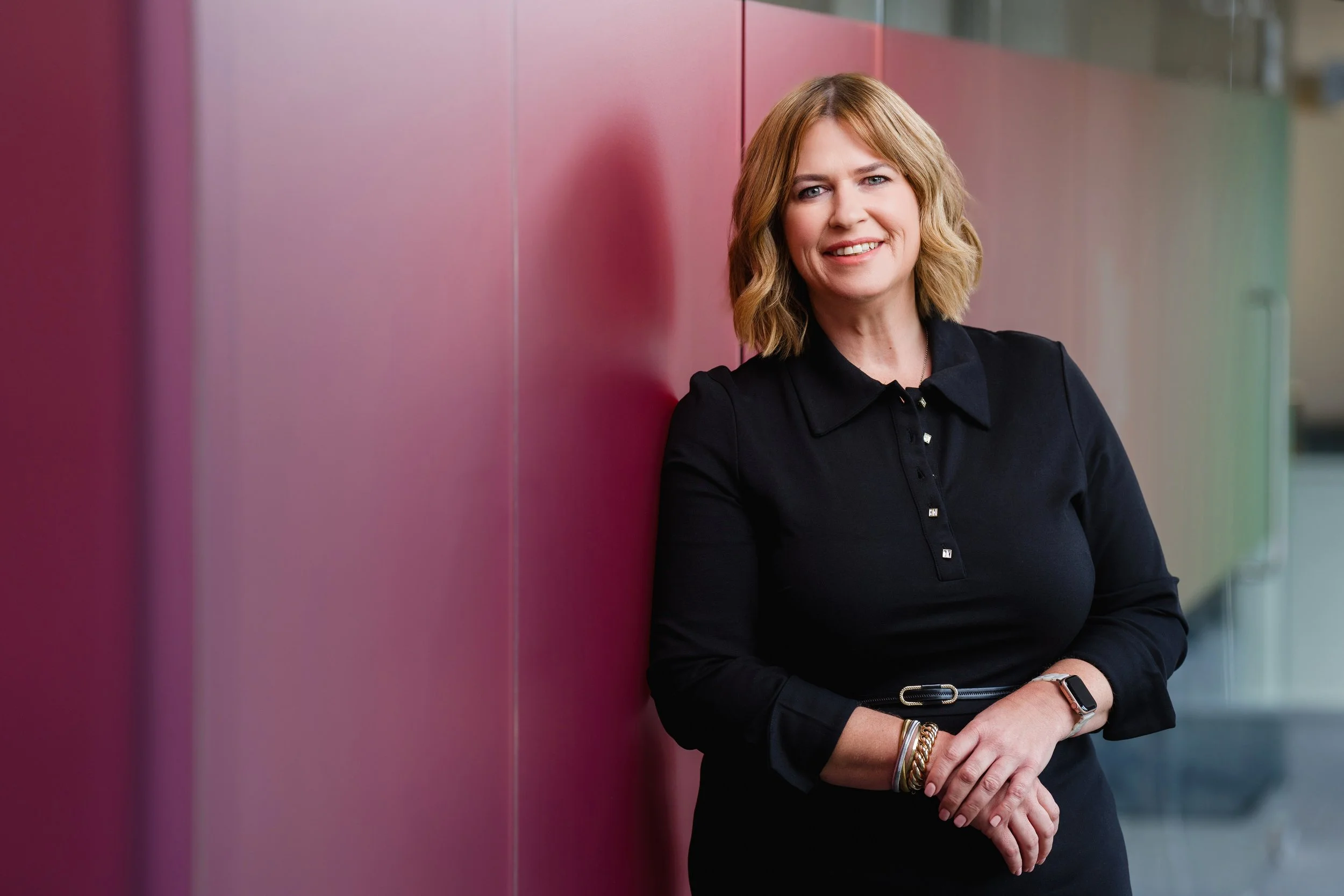 A woman with blonde hair wearing a black dress, standing and smiling while leaning against a colorful wall.
