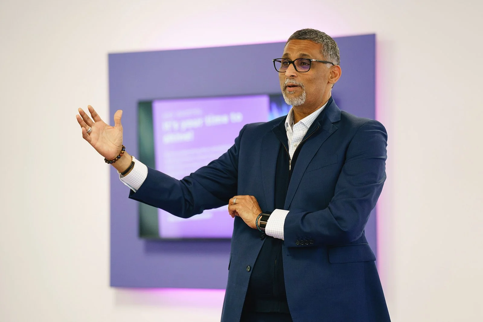 A man in a dark suit and glasses giving a presentation in front of a purple and white background.