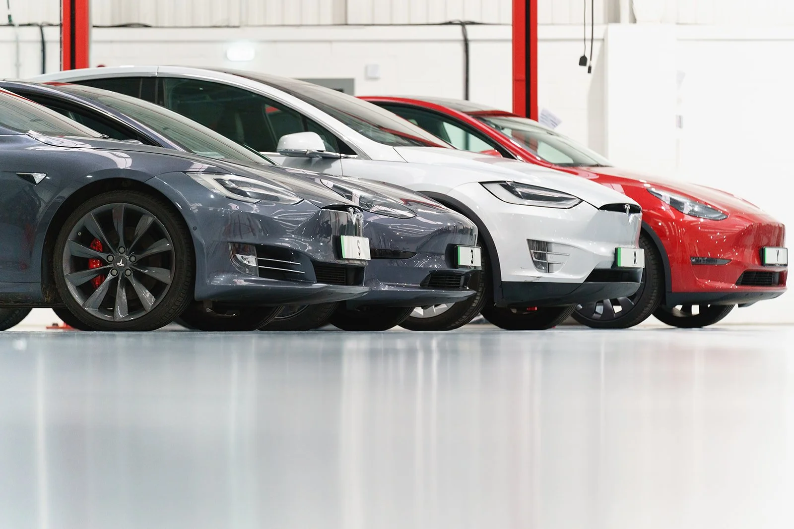 A row of four Tesla cars parked indoors, including a dark gray Model S, a white Model X, and two red Model 3s.