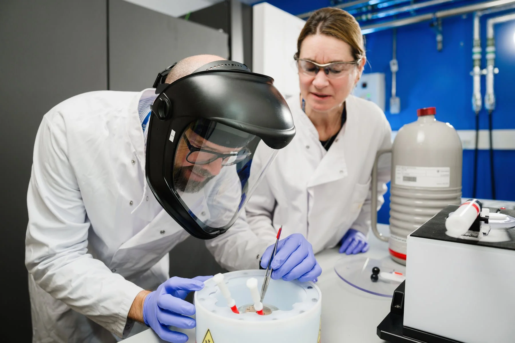 Two scientists in lab coats conducting an experiment, one wearing a protective face shield and gloves, working with a centrifuge or similar lab equipment, while the other watches attentively in a laboratory setting.