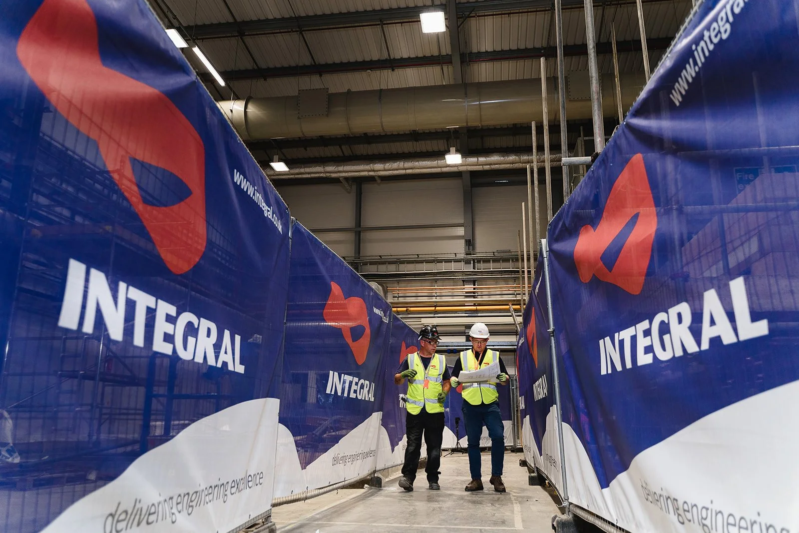 Two construction workers wearing helmets and high-visibility vests standing and reviewing blueprints inside an industrial building surrounded by large blue banners with the word 'INTEGRAL' and a red logo, with pipes visible on the ceiling.
