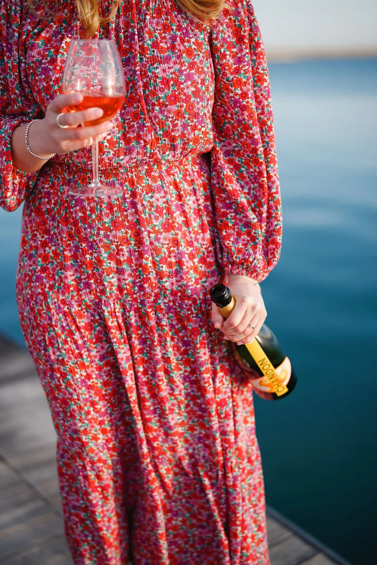 A person in a long, colorful floral dress holding a glass of rosé wine and a bottle of champagne standing on a wooden dock near water.