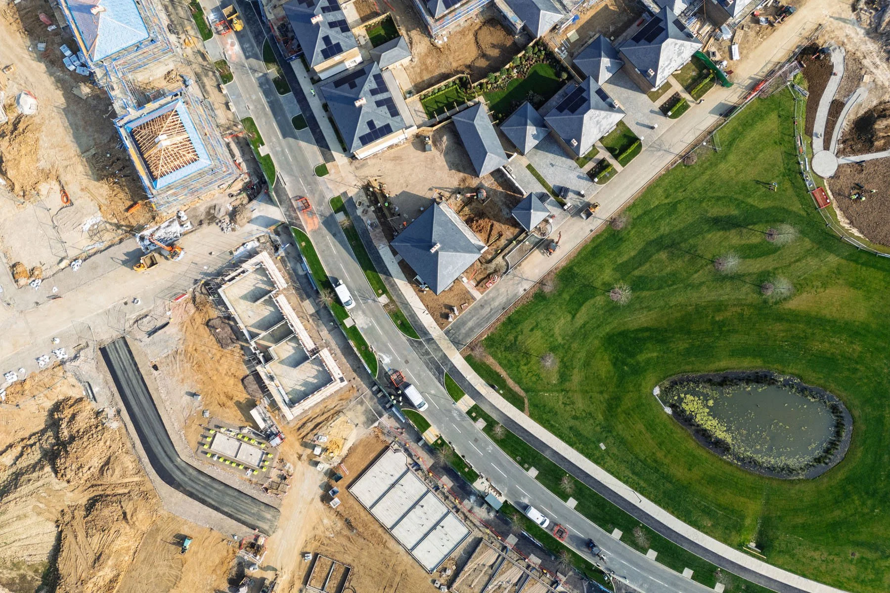 Aerial view of a residential area under construction with houses being built, a small pond, and a green park area.