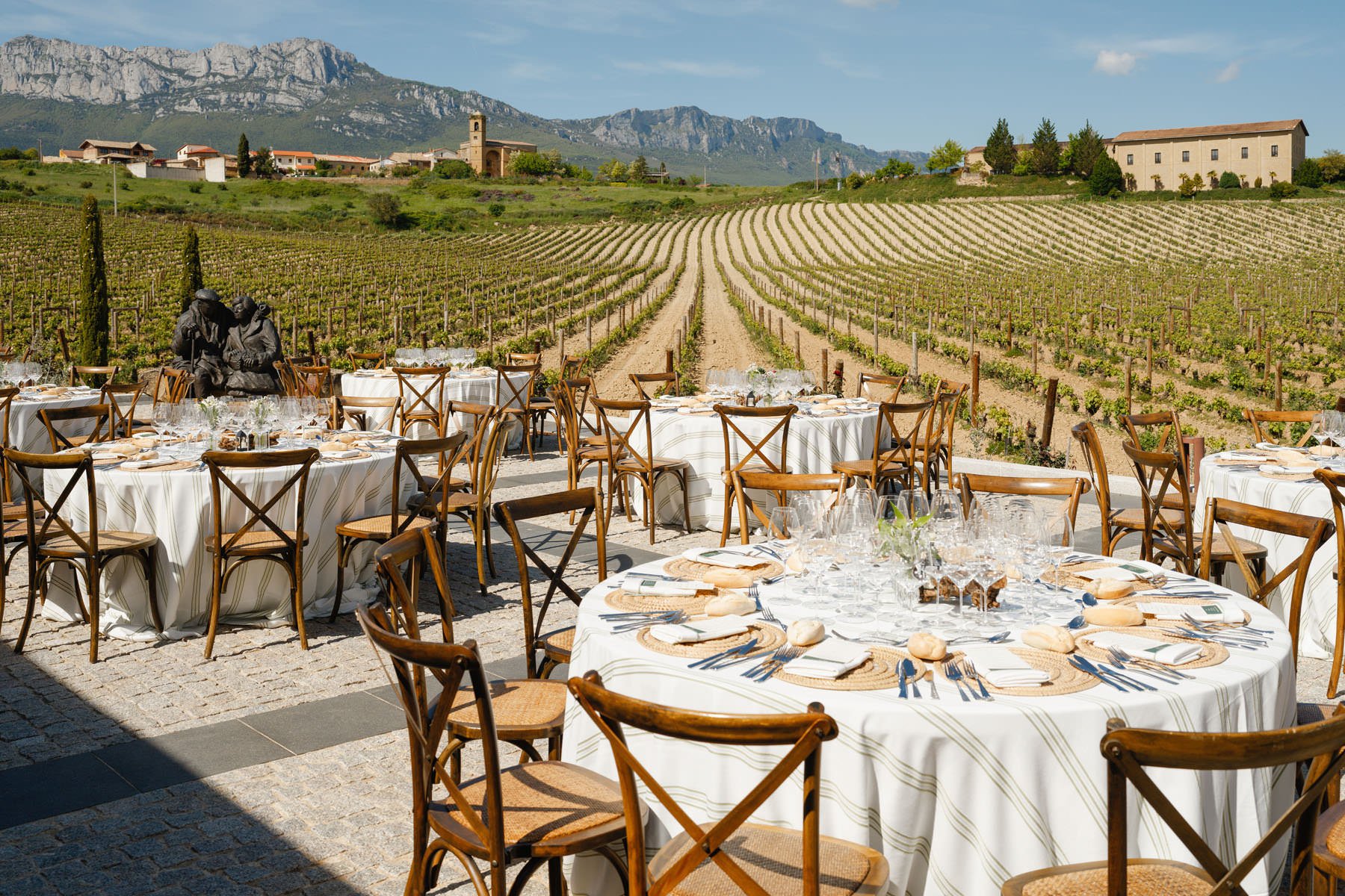 Outdoor dining setup with round tables covered in white tablecloths and wooden chairs, set against a backdrop of a vineyard with mountains and a small village in the distance under a partly cloudy sky.