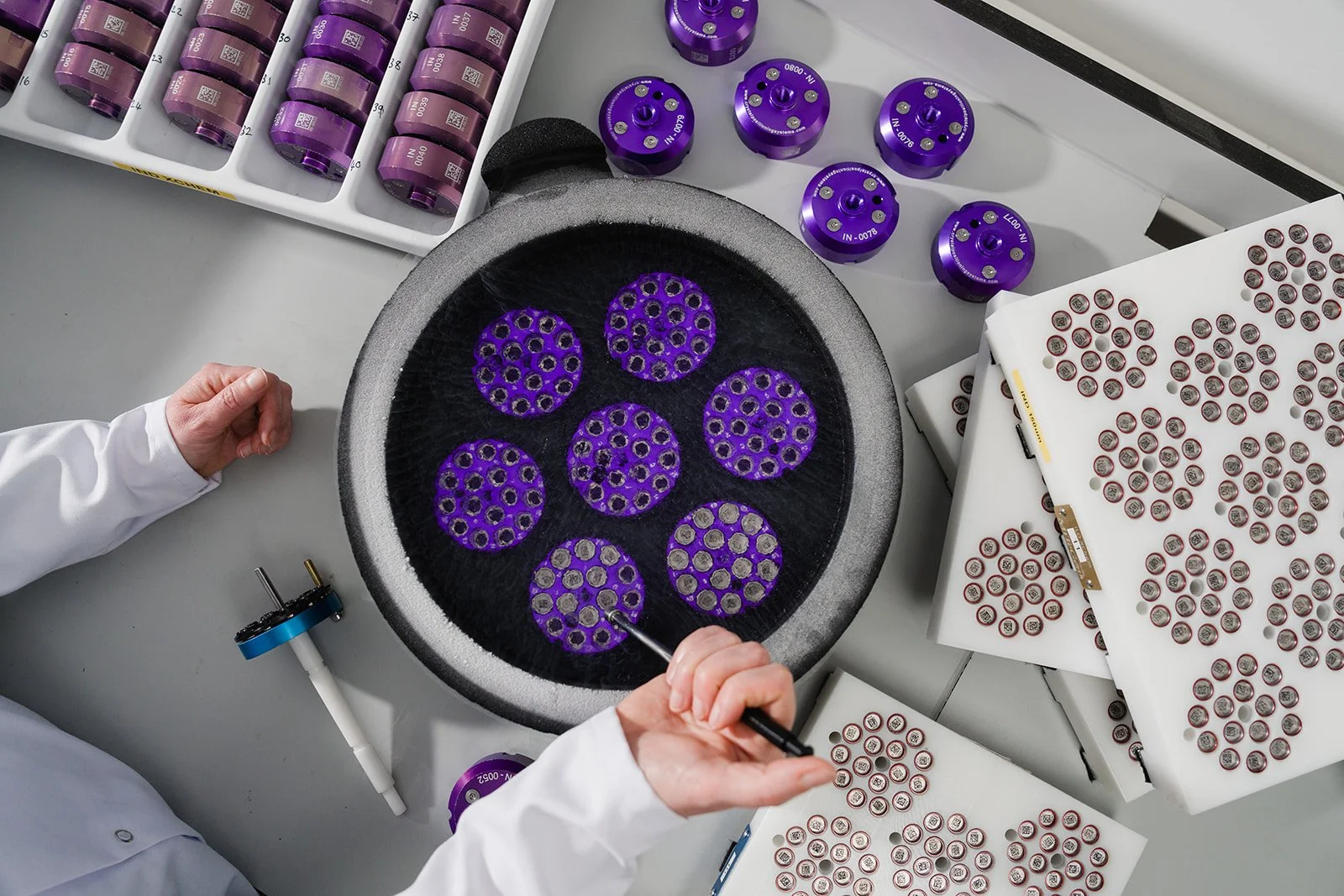 A scientist working with purple and red cylindrical components and small metallic parts on a work bench, possibly engaged in a manufacturing or assembly process of medical or electronic devices.