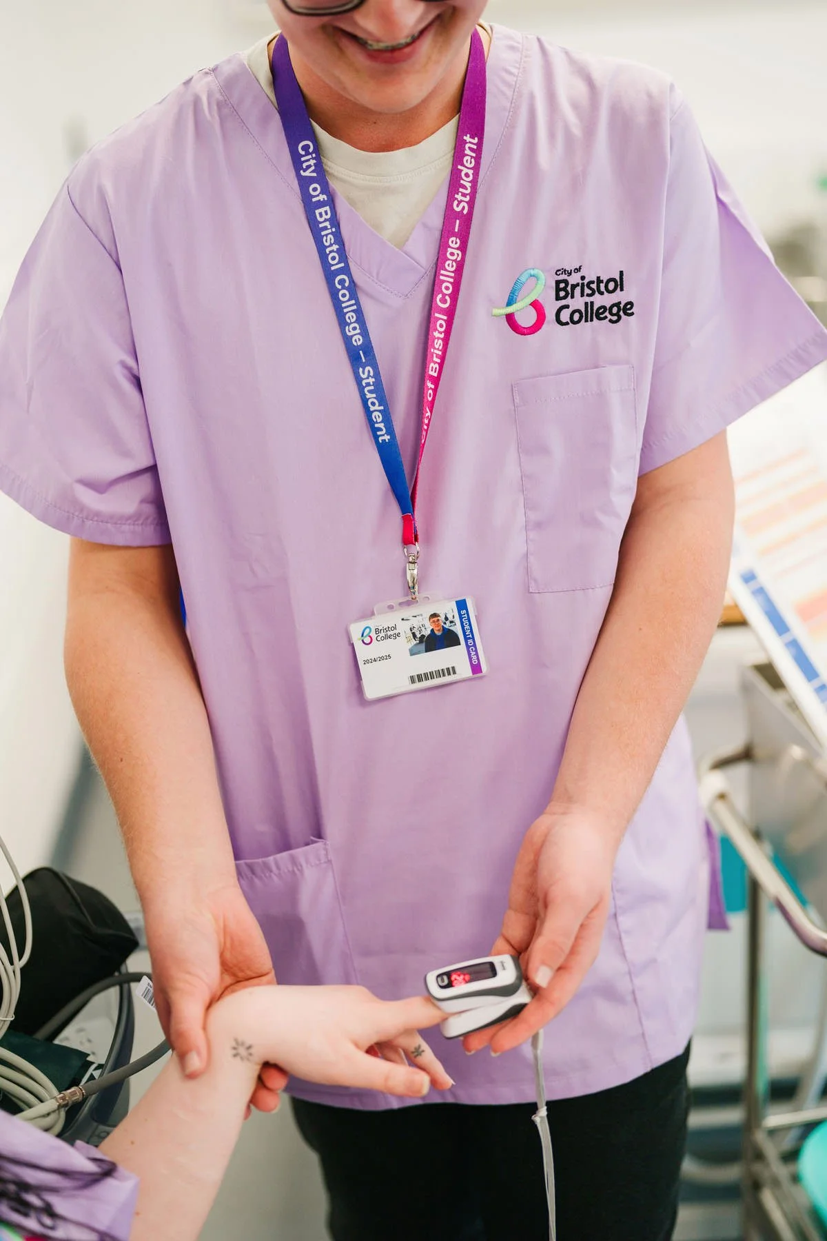 A healthcare worker in a purple scrub top with a Bristol College badge, taking a patient's pulse with a pulse oximeter. The worker is smiling, and the patient's hand is visible with some tattoos.