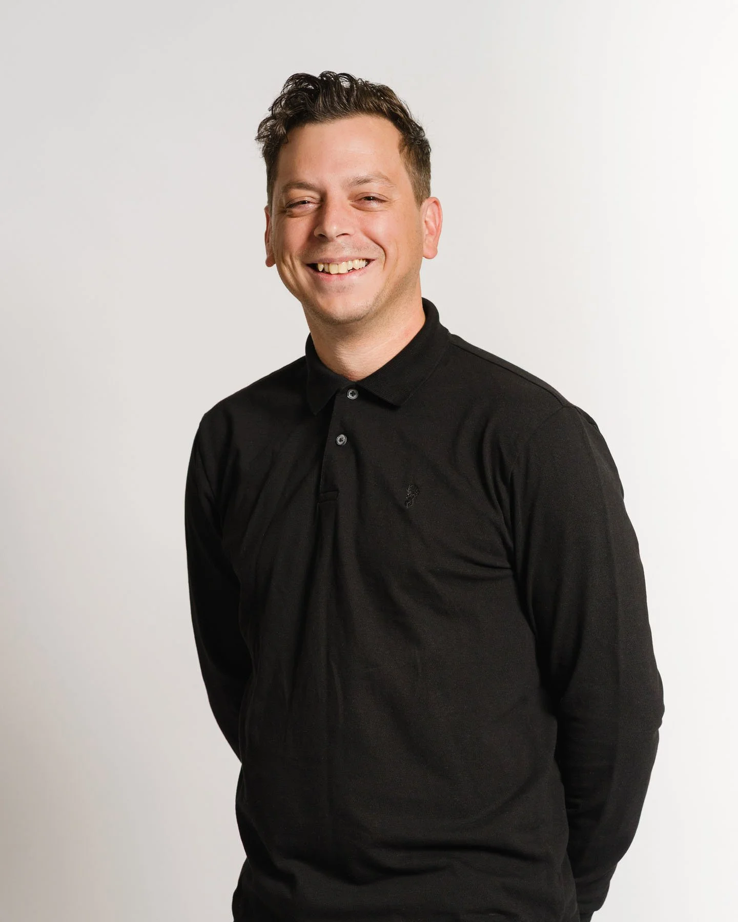 A smiling man in a black collared shirt standing against a plain white background.