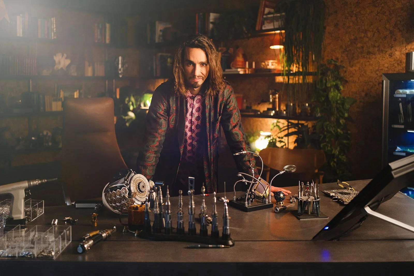 A man with long hair leaning on a table with various electronic devices and tools in a warmly lit room with bookshelves and plants.