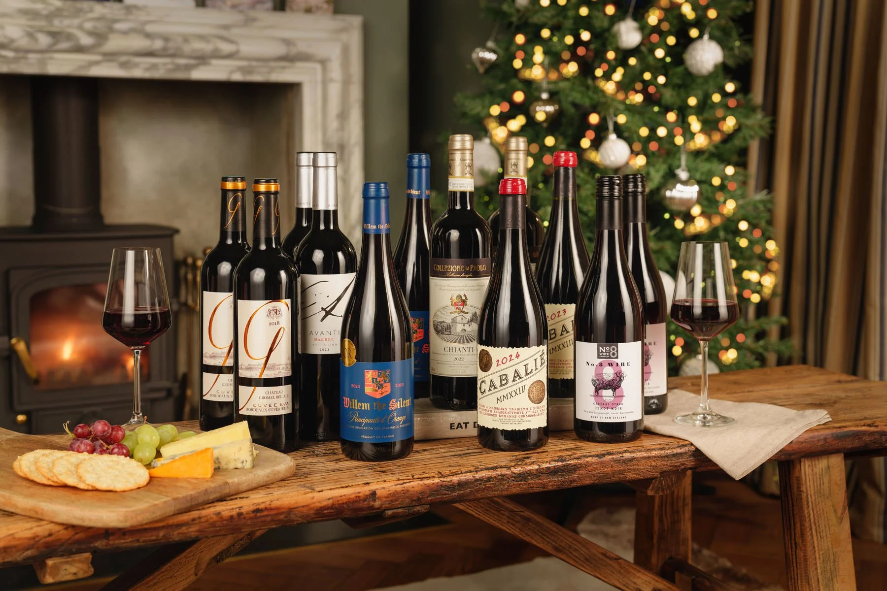 A wooden table with multiple bottles of red wine, two glasses of red wine, a cheese platter with grapes, crackers, and cheese, in front of a decorated Christmas tree with lights and ornaments.