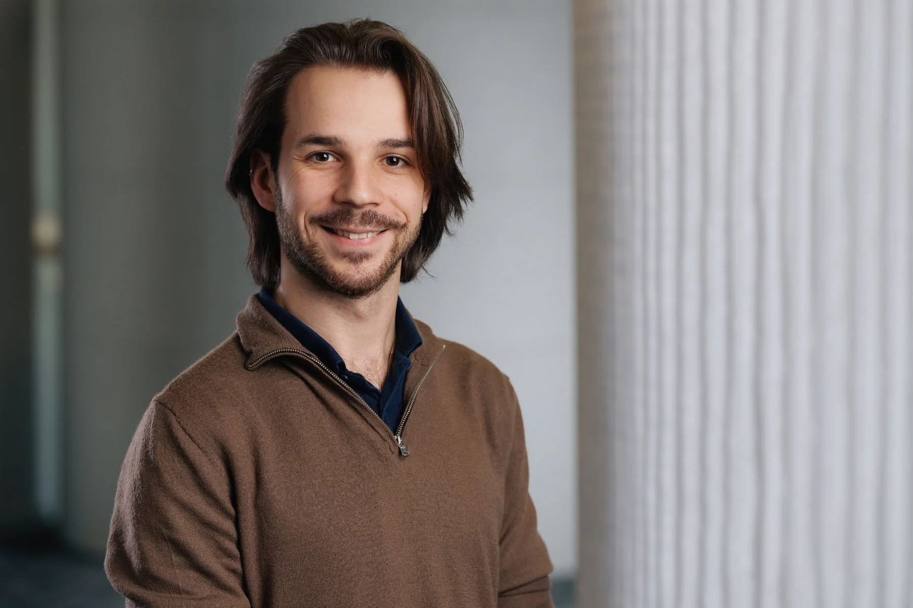A man with medium-length brown hair and facial hair smiling, wearing a brown sweater over a dark collared shirt, standing indoors near a large window with vertical blinds.