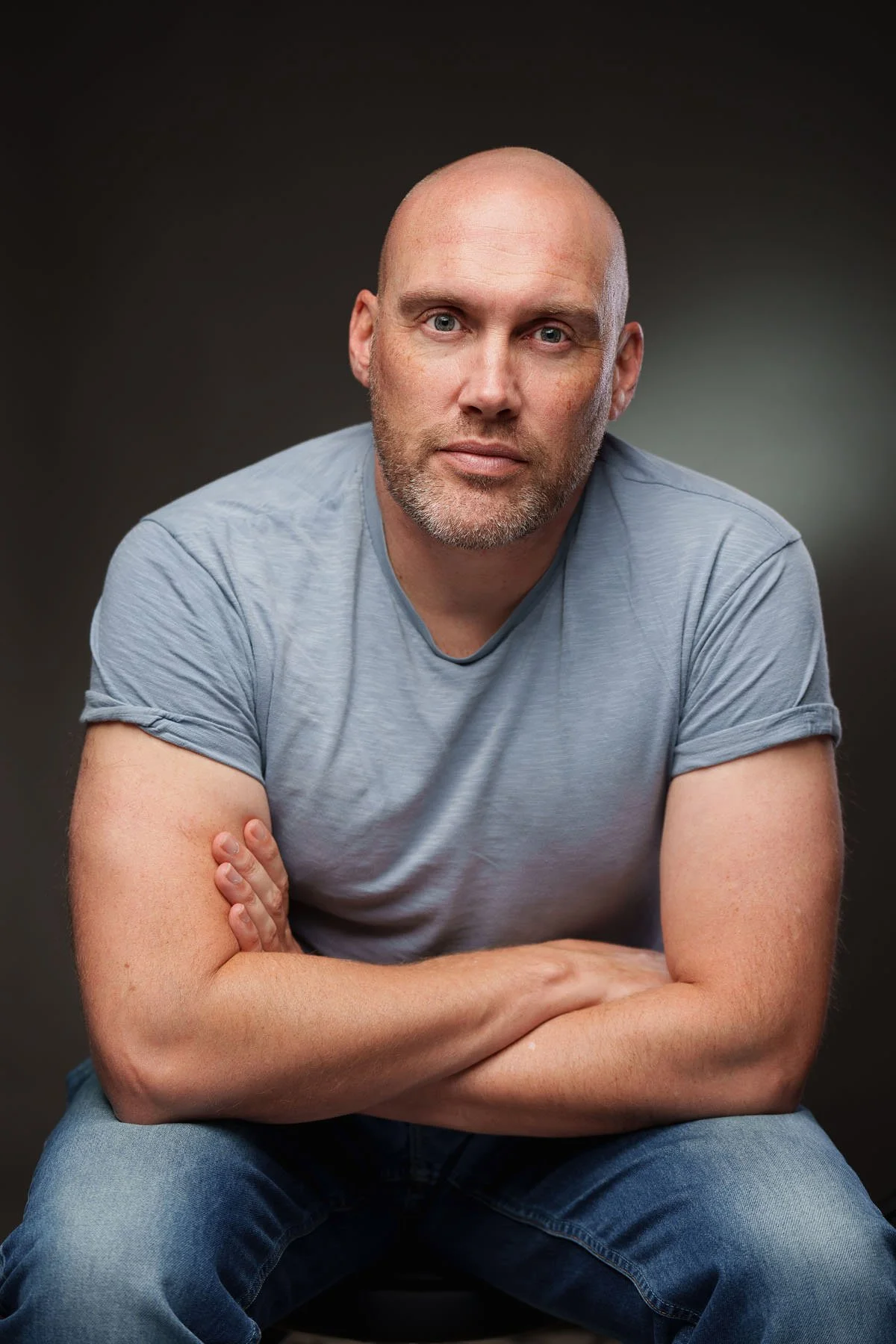 A man with a shaved head and a short beard, wearing a light gray t-shirt and jeans, sitting with his arms crossed, looking at the camera against a dark gradient background.