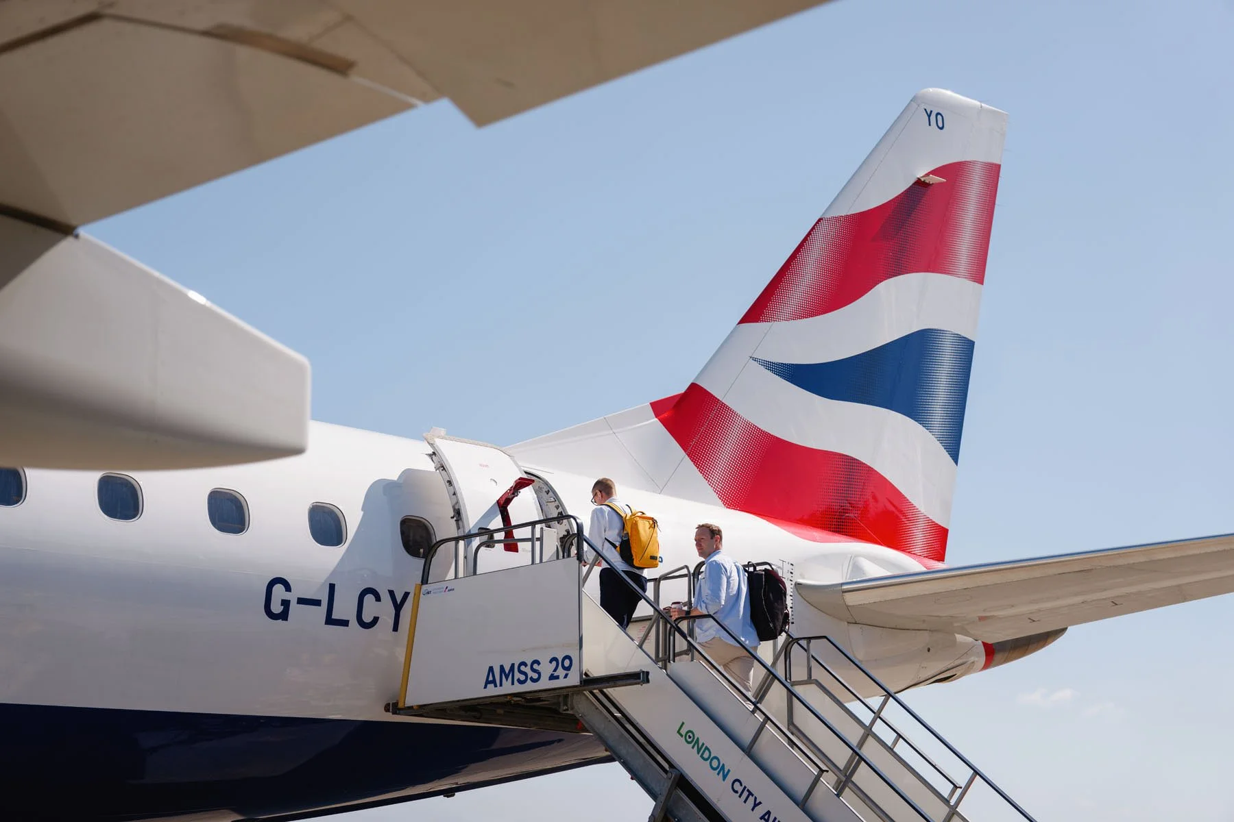 Two passengers boarding a British Airways aircraft via stairs, with the aircraft's tail showing a red, white, and blue design, against a clear blue sky.