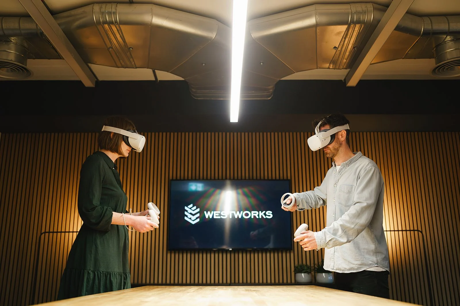 Two people wearing white virtual reality headsets and holding controllers in a modern room with wooden wall paneling and a large screen displaying the Westworks logo.
