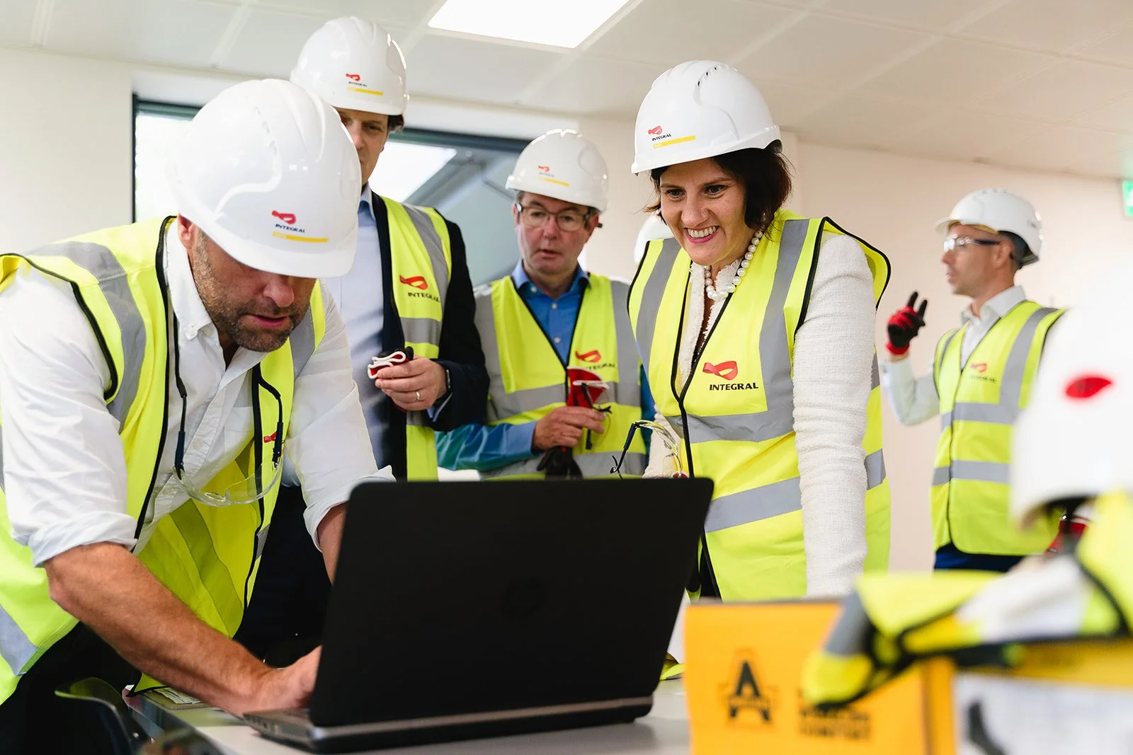 Group of engineers and architects wearing yellow safety vests and white hard hats discussing plans around a laptop in an indoor setting.