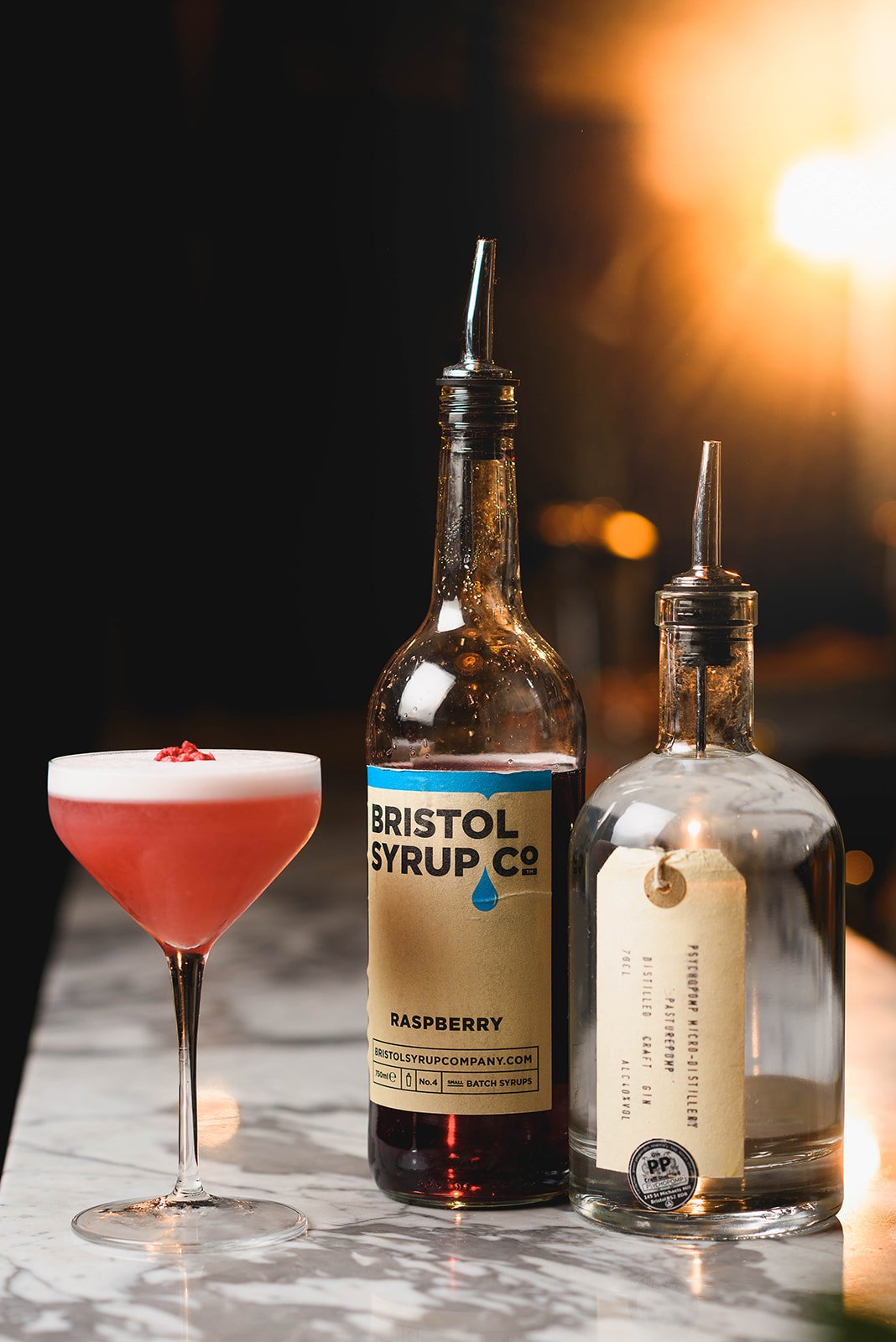 A pink cocktail in a martini glass with a red garnish, a bottle of Bristol Syrup Co. raspberry syrup, and a clear bottle with a spout on a marble surface with warm background lighting.