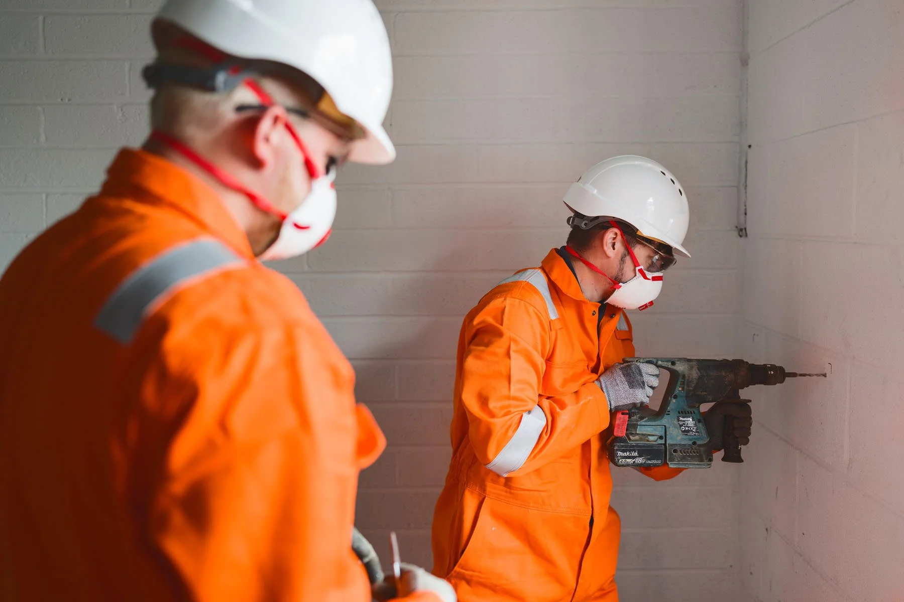 Two construction workers wearing orange jumpsuits, white safety helmets, goggles, gloves, and respiratory masks working inside a building with white brick walls. One worker is using a blue power drill against the wall, while the other supervises.