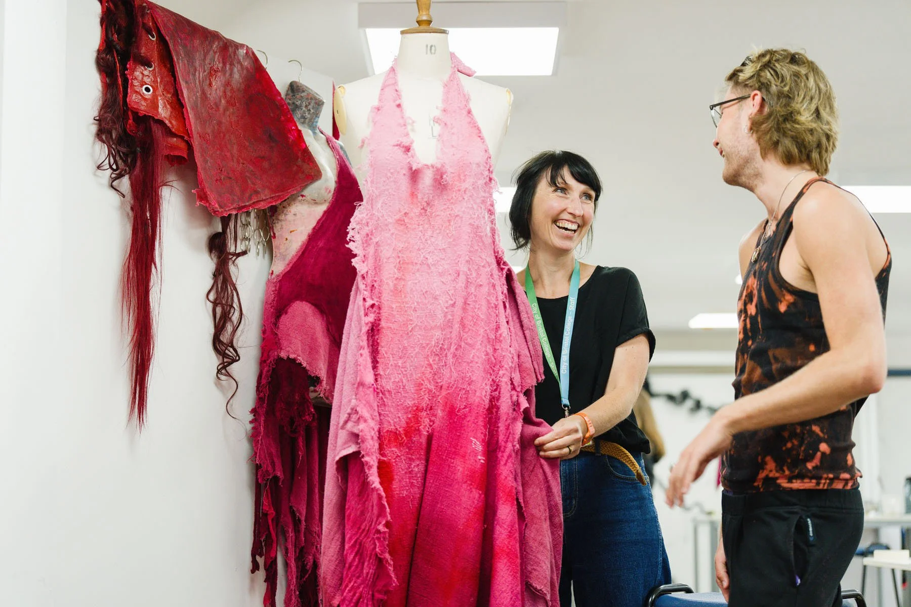 Two people smiling and talking near a mannequin dressed in a pink, textured garment in a bright, well-lit room. There are additional fabric swatches hanging on the wall.