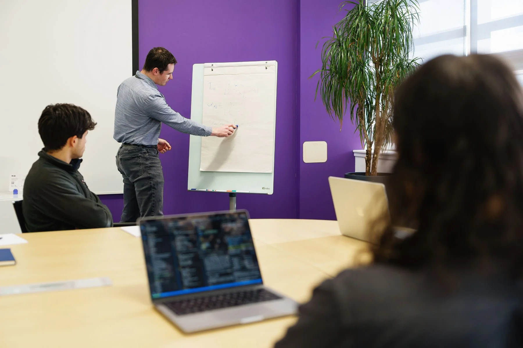 A man presents in front of two colleagues in a meeting room, with a whiteboard and purple accent wall, while others observe, one working on a laptop.