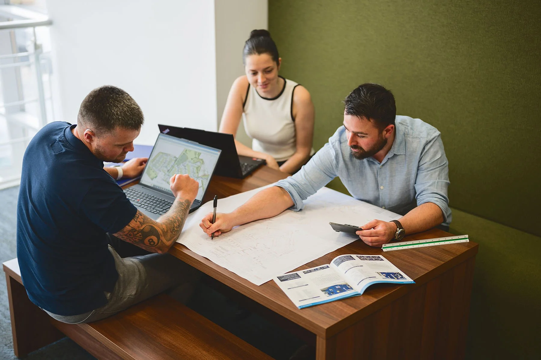 Three people working on maps and digital devices at a wooden table in a bright room.