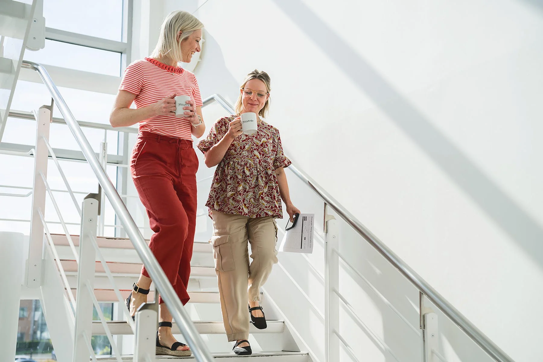 Two women with glasses and light hair walking down a staircase with white walls and large windows. They are holding mugs and a piece of paper, wearing casual clothing and sandals.