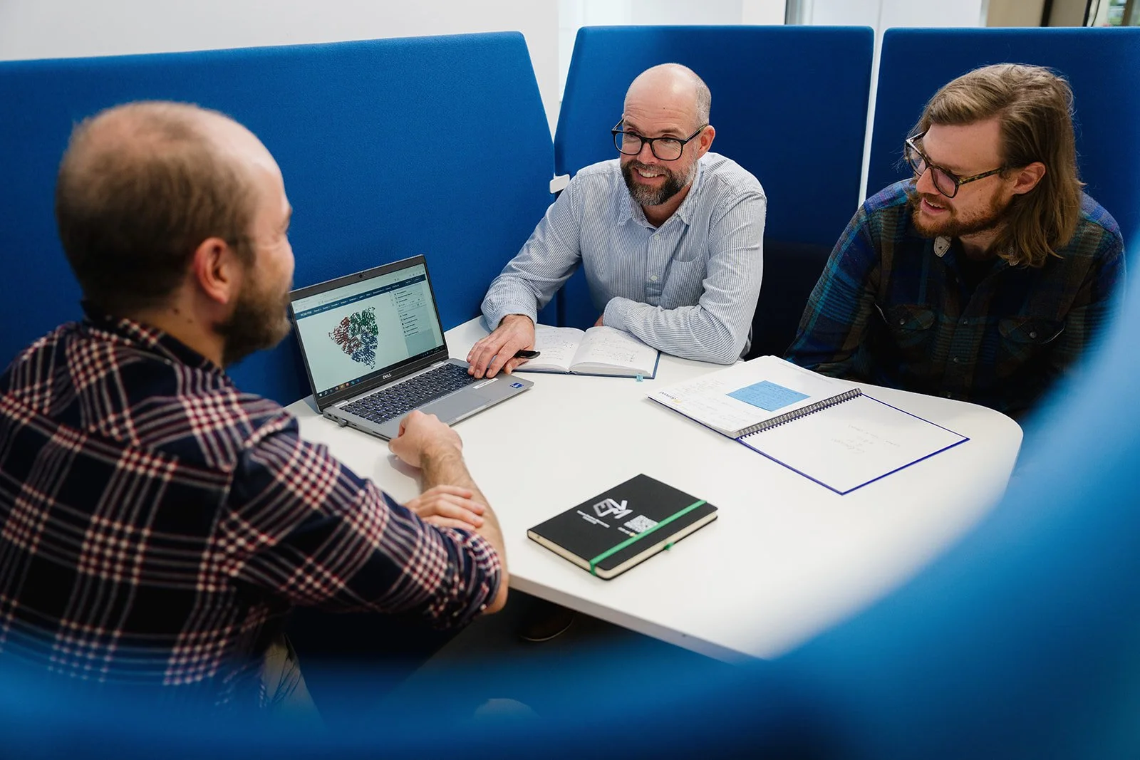 Three men sitting at a white table in a meeting room, engaged in a discussion, with a laptop displaying a colorful diagram, notebooks, and pens on the table.