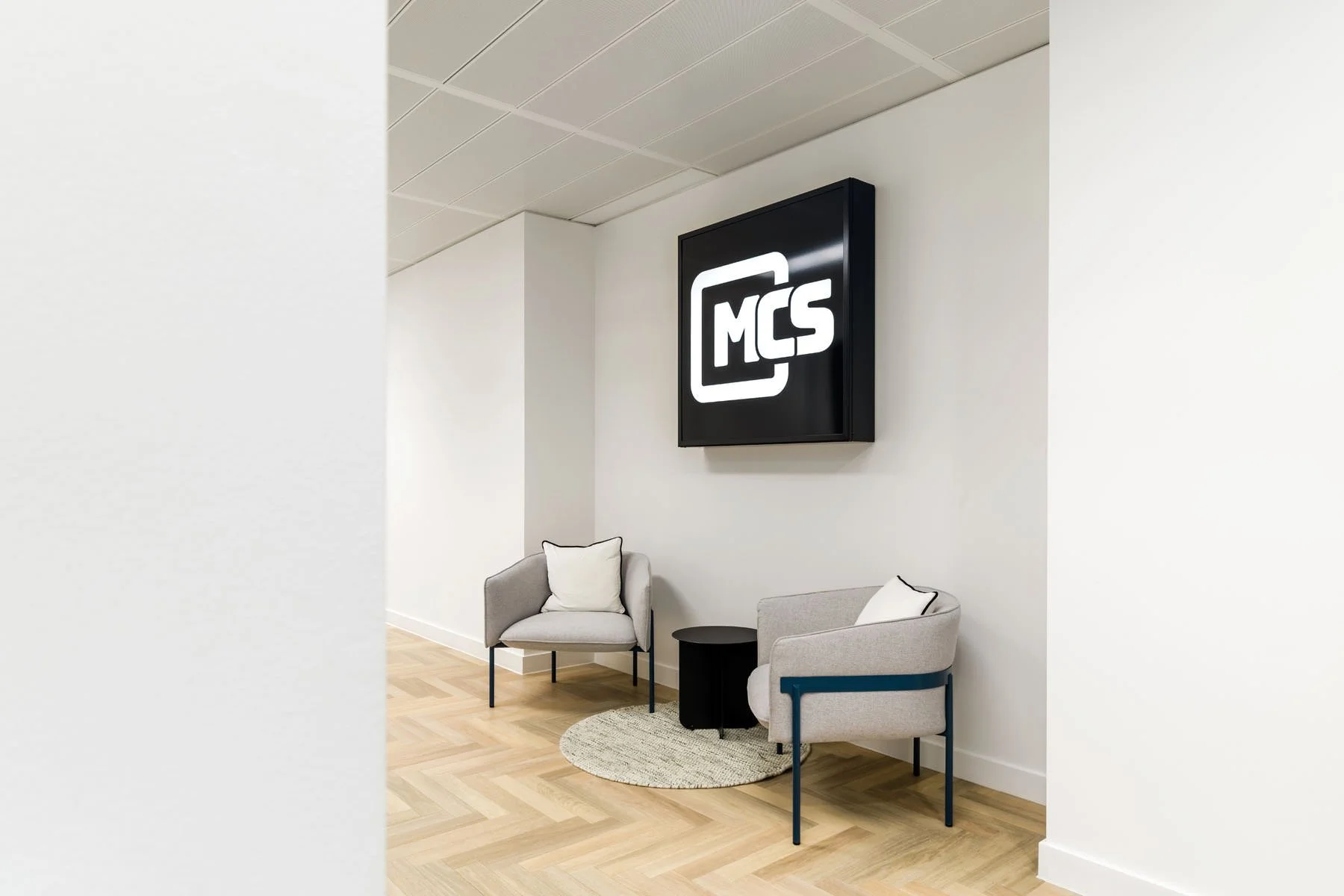 Waiting area with two light gray armchairs, a small black round table, a round beige rug, and a black framed sign on a white wall with the letters "MCS" in white inside a black square.