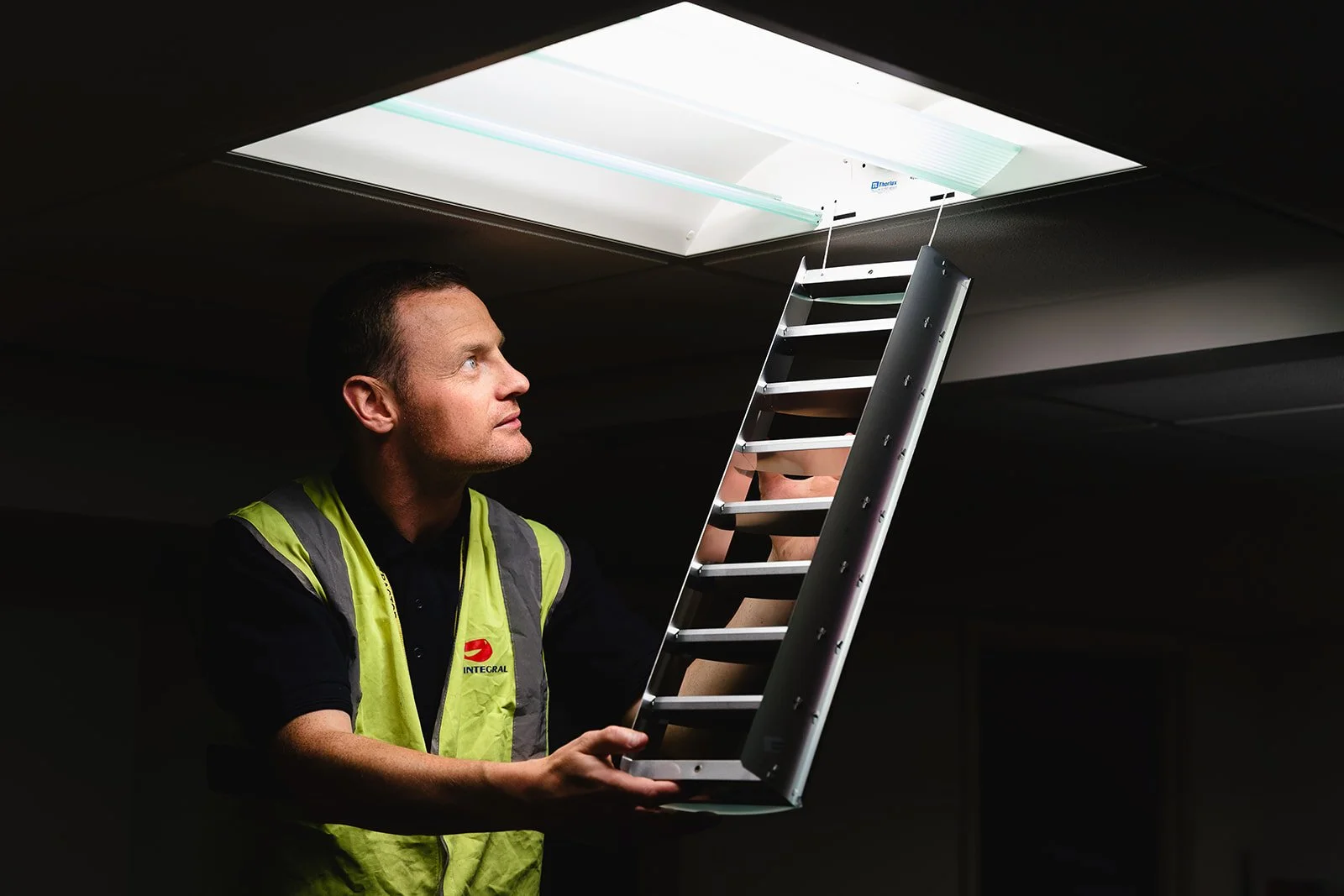 A man wearing a yellow safety vest is holding a small ladder, about to climb into an open roof skylight in a ceiling.