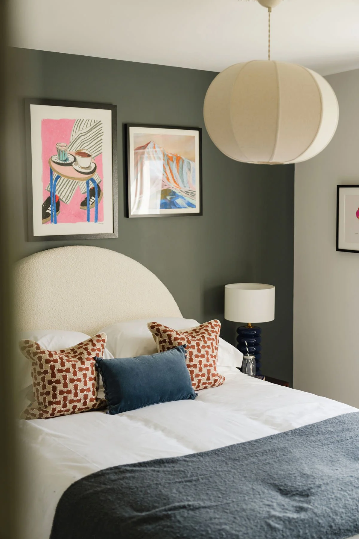 A cozy bedroom with a bed, decorative pillows, a white headboard, wall art, a table lamp, and a large fabric pendant light.