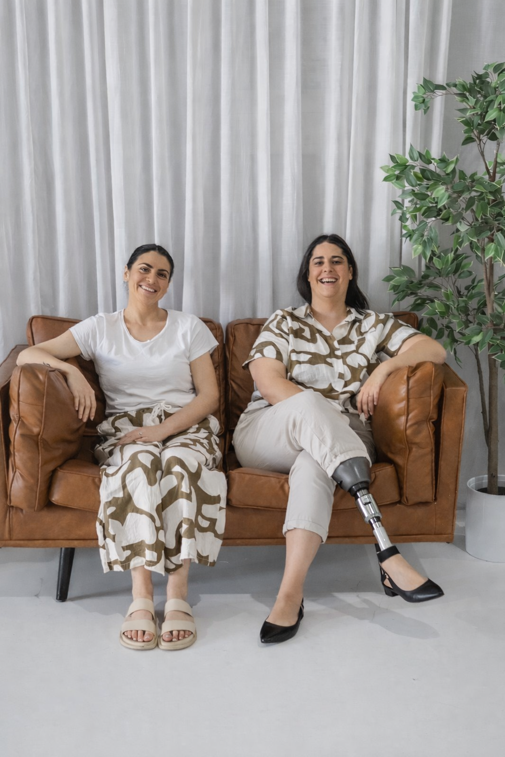 Two women sitting on a brown leather couch, smiling at the camera, with a white curtain backdrop and a green potted plant on the right.
