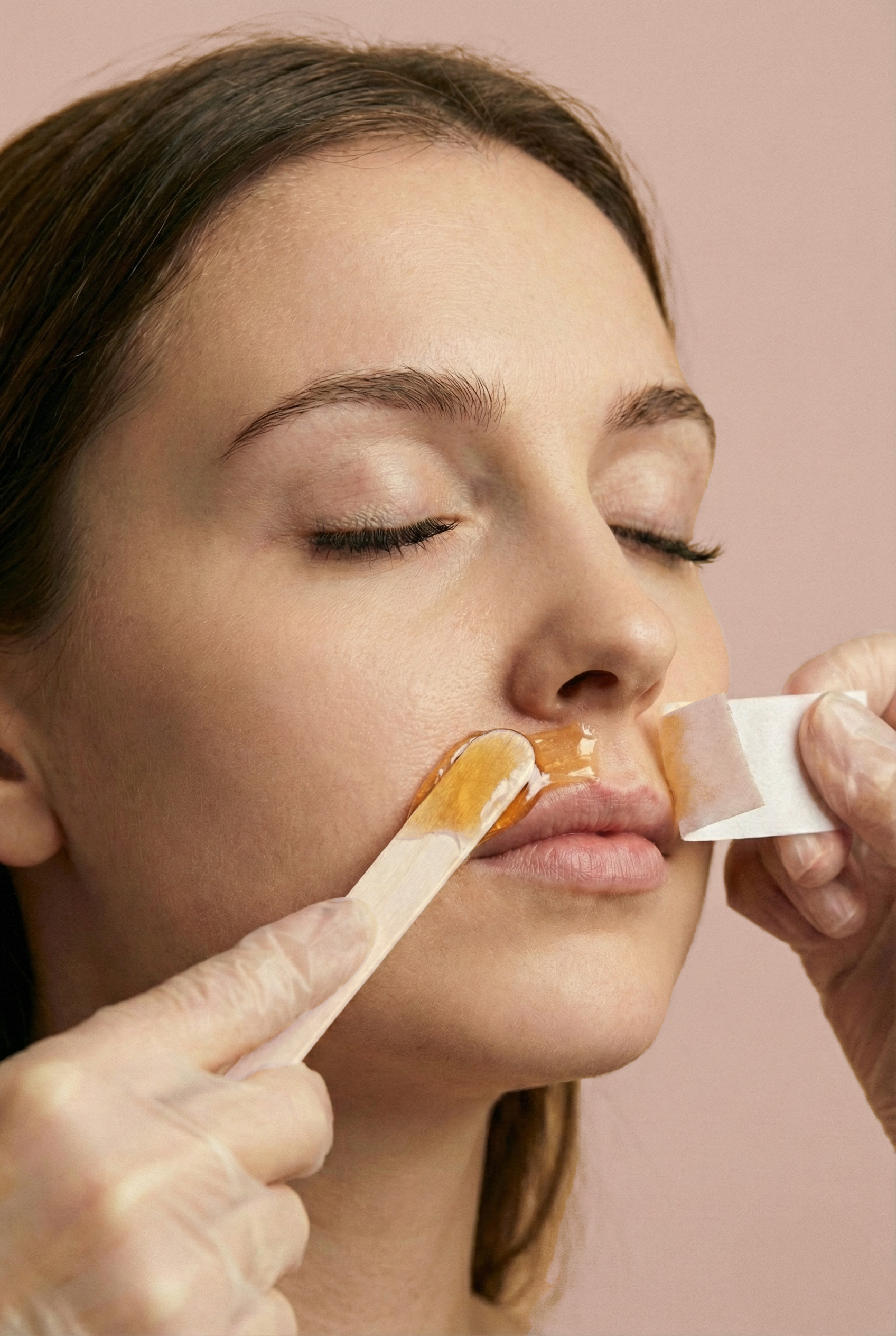 Close-up of a woman with closed eyes receiving a lip treatment from a professional using a spatula and wax strips, with a pink background.