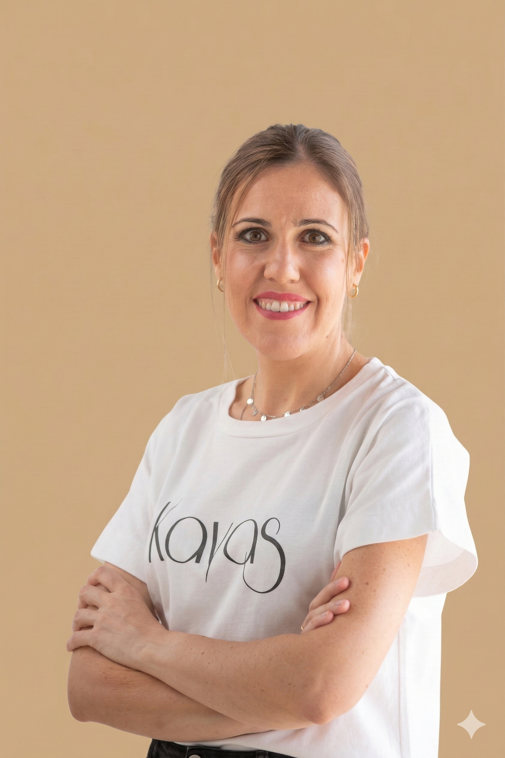 A smiling woman with light brown hair tied back, wearing a white t-shirt with the word 'kayas' printed on it, standing with arms crossed against a plain beige background.