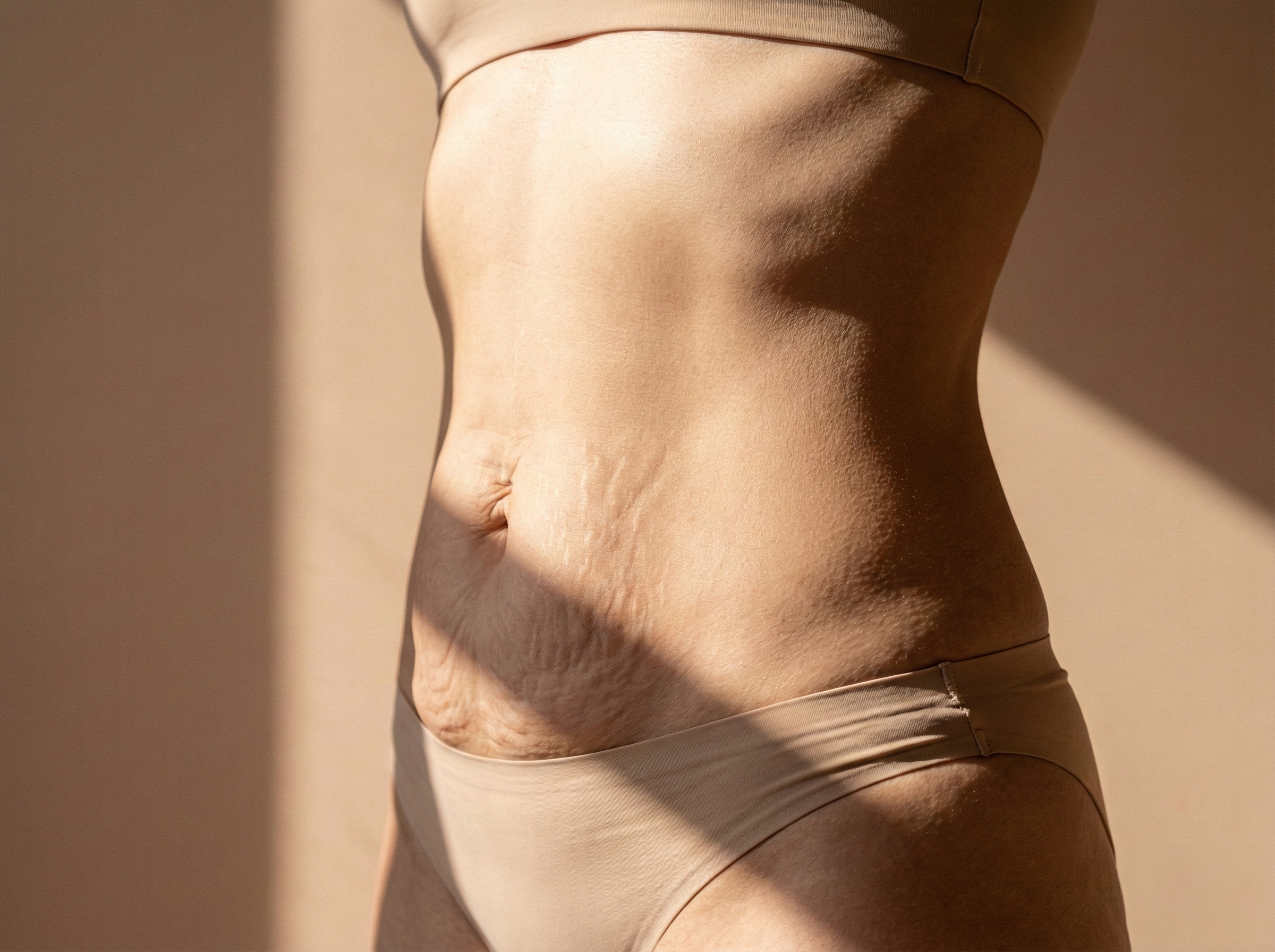 Close-up of a woman's midsection with toned abs and a navel, wearing beige underwear, with a shadow cast on her side.
