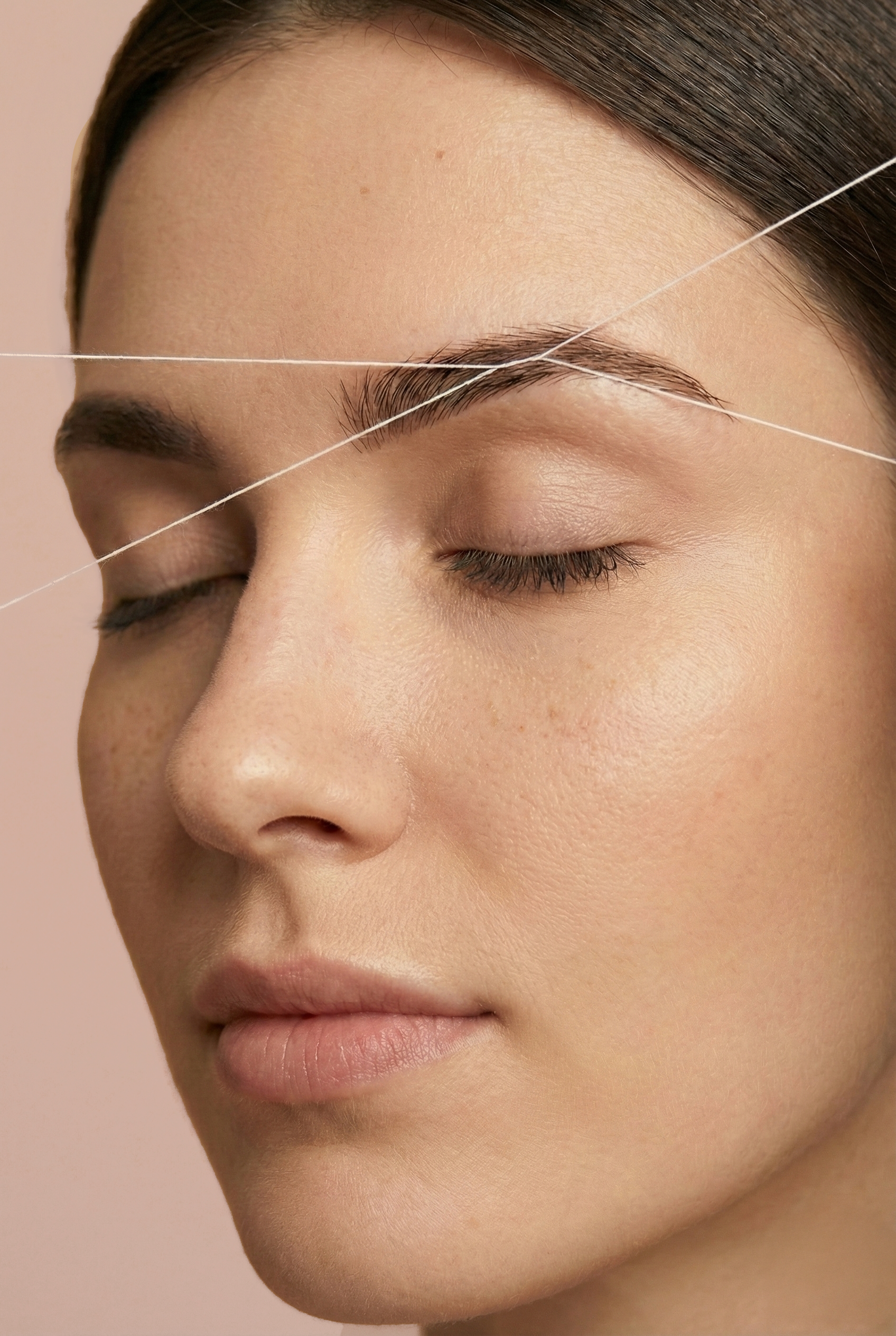 Close-up of a woman's face with closed eyes and smooth skin, with two pieces of white thread stretched across her face.