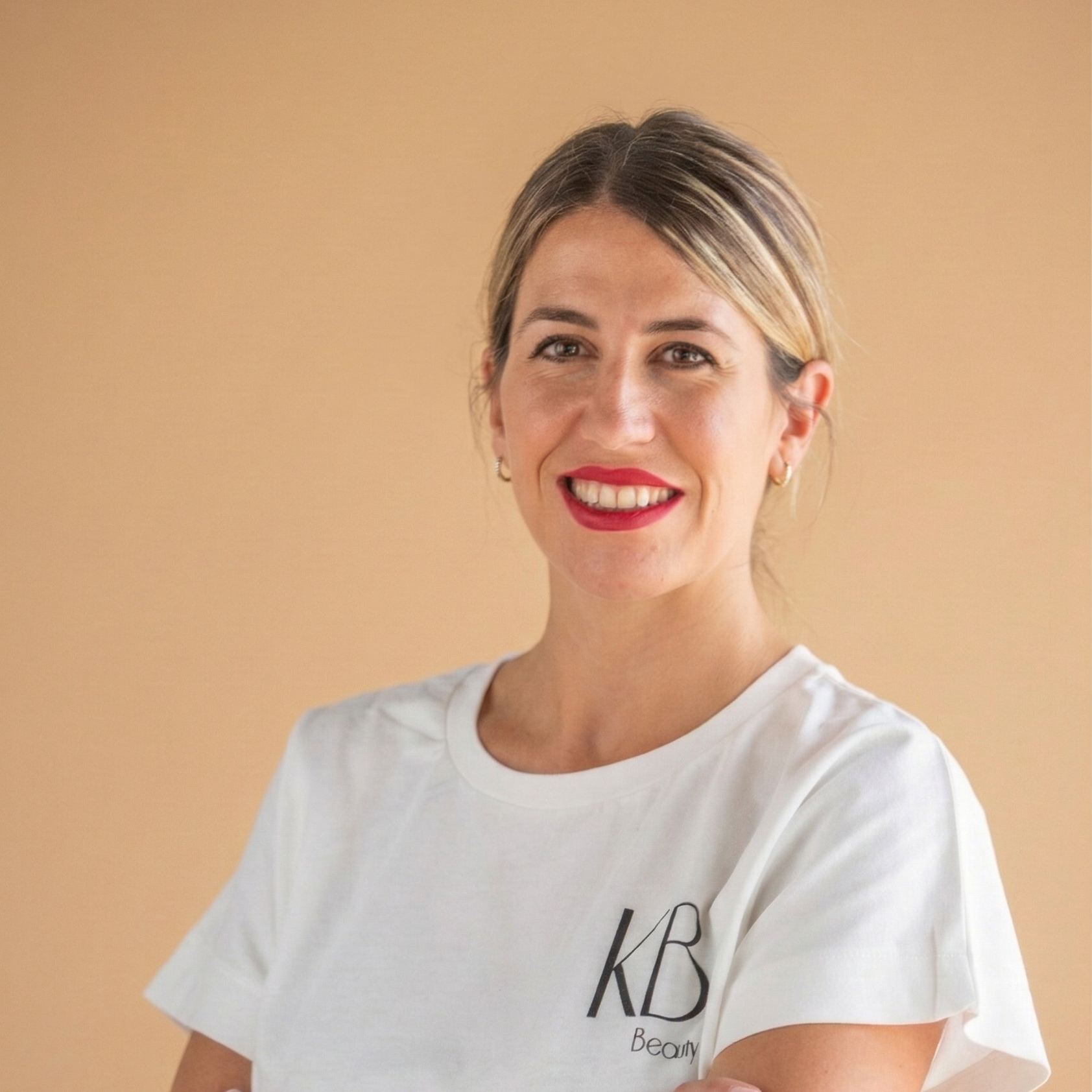 A woman with light skin, brown hair with blonde highlights, smiling, wearing red lipstick and small hoop earrings, during daytime, in a white T-shirt with black text that says 'KB Beauty'.