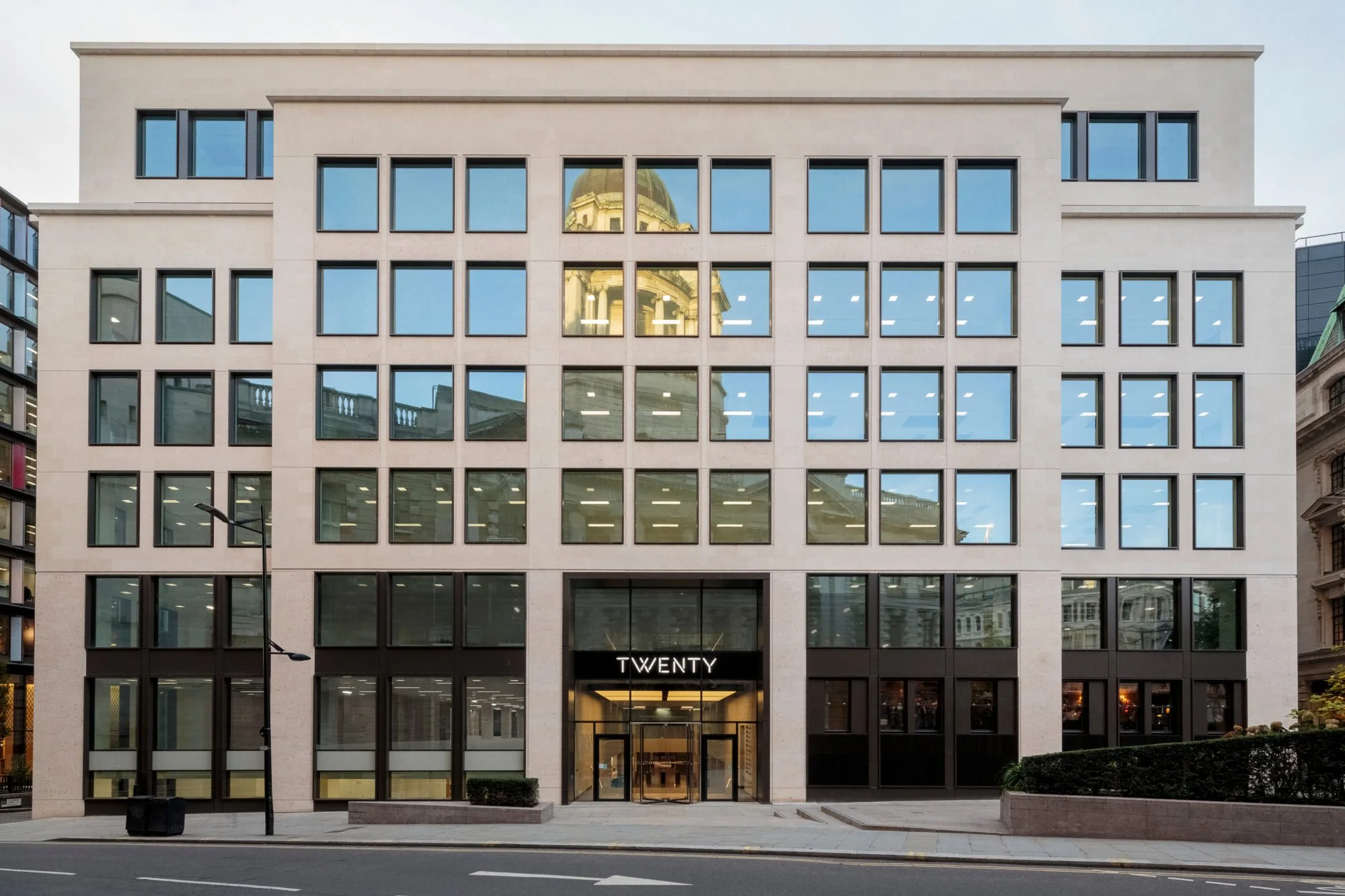 Modern multi-story office building with large glass windows reflecting a dome building, and the sign 'TWENTY' above the entrance, on a city street.
