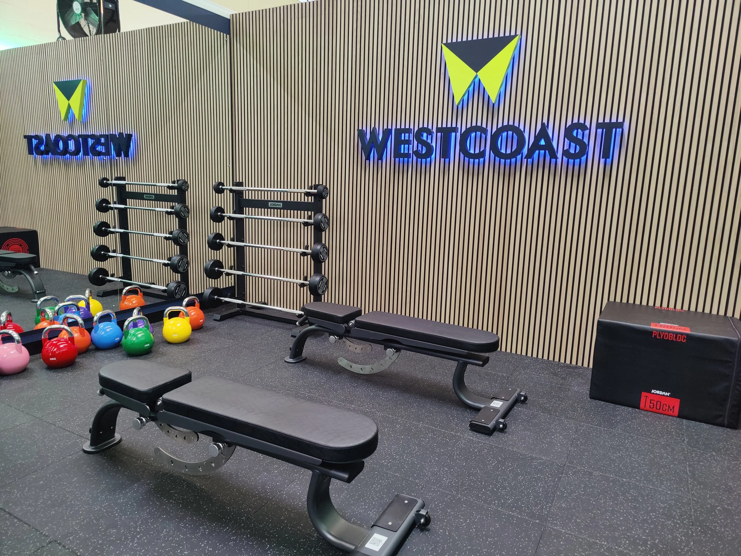 Workout area featuring two black workout benches, dumbbell racks with dumbbells, colorful kettlebells and a wall with a large blue sign reading ‘WEST COAST’ and their logo.