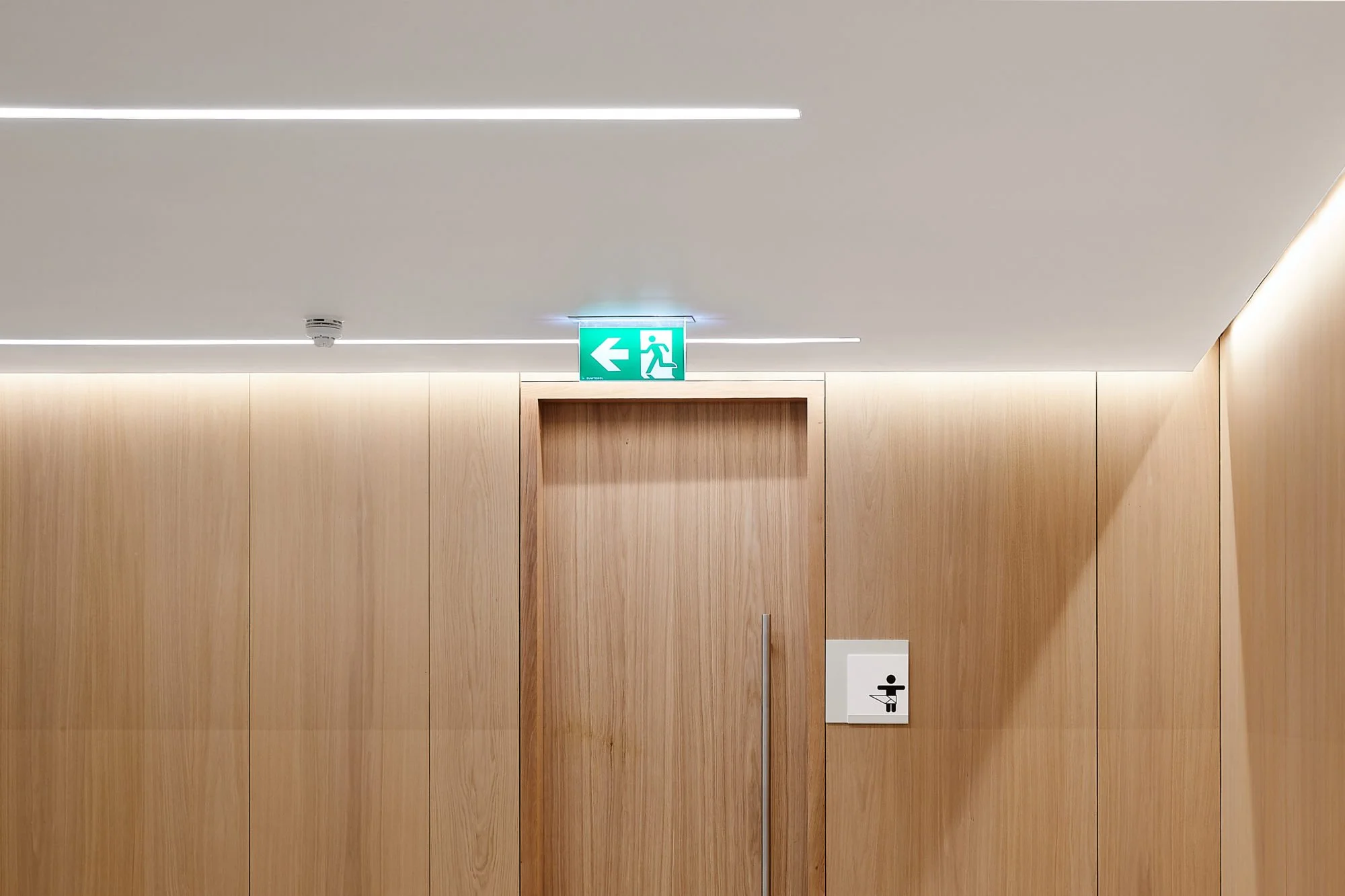 Wood-paneled wall with a closed door, an emergency exit sign above it pointing to the left, and a white button with a black icon of a person washing their hands.