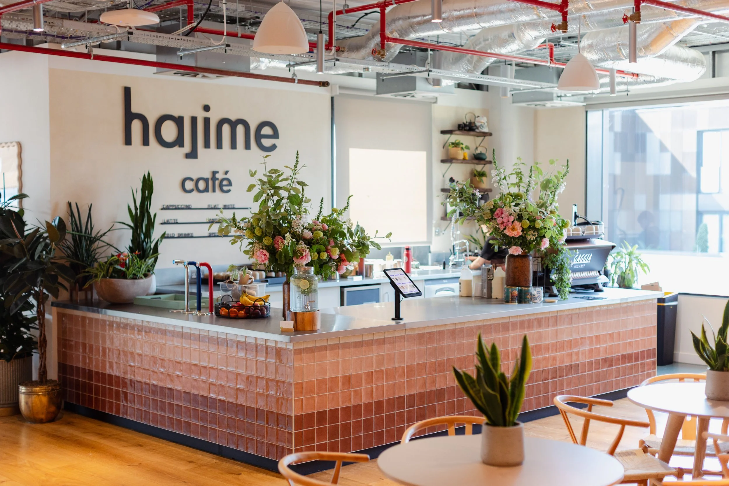 Interior of a modern café with a pink tiled counter, large floral arrangements, potted plants, and a sign that reads 'hajime café' on the wall.