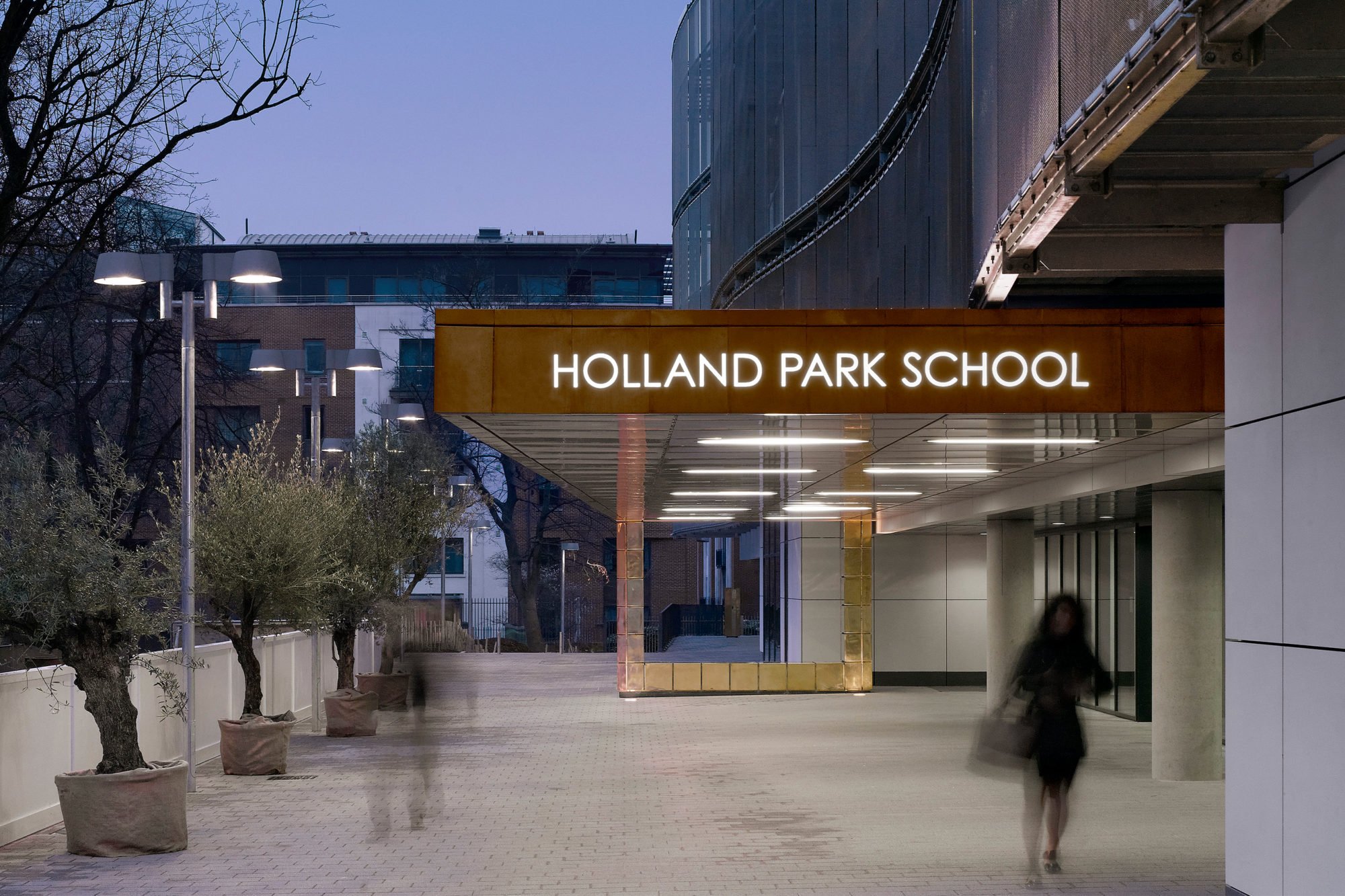 The entrance to Holland Park School in an urban setting during dusk with a canopy featuring illuminated sign and modern architecture. A woman is walking past, with other blurred figures nearby.