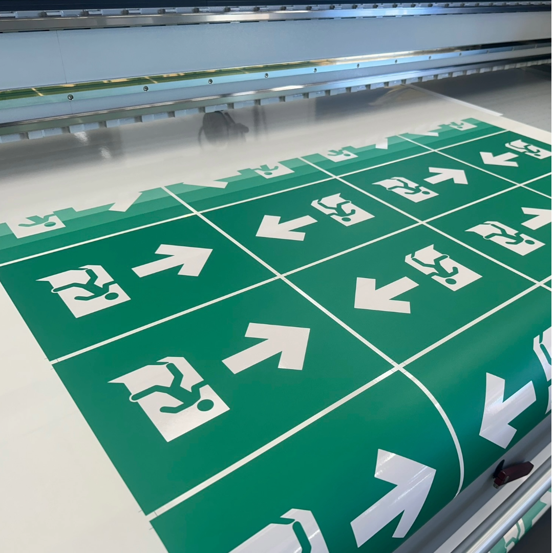 A printing press printing green emergency exit signs with white directional arrows and running figure symbols.