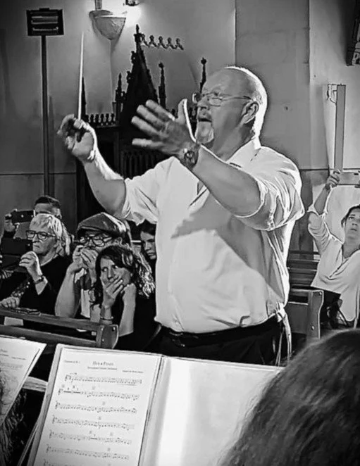 Un chef d'orchestre dirige un groupe de musiciens dans une salle de concert, avec des spectateurs en arrière-plan.